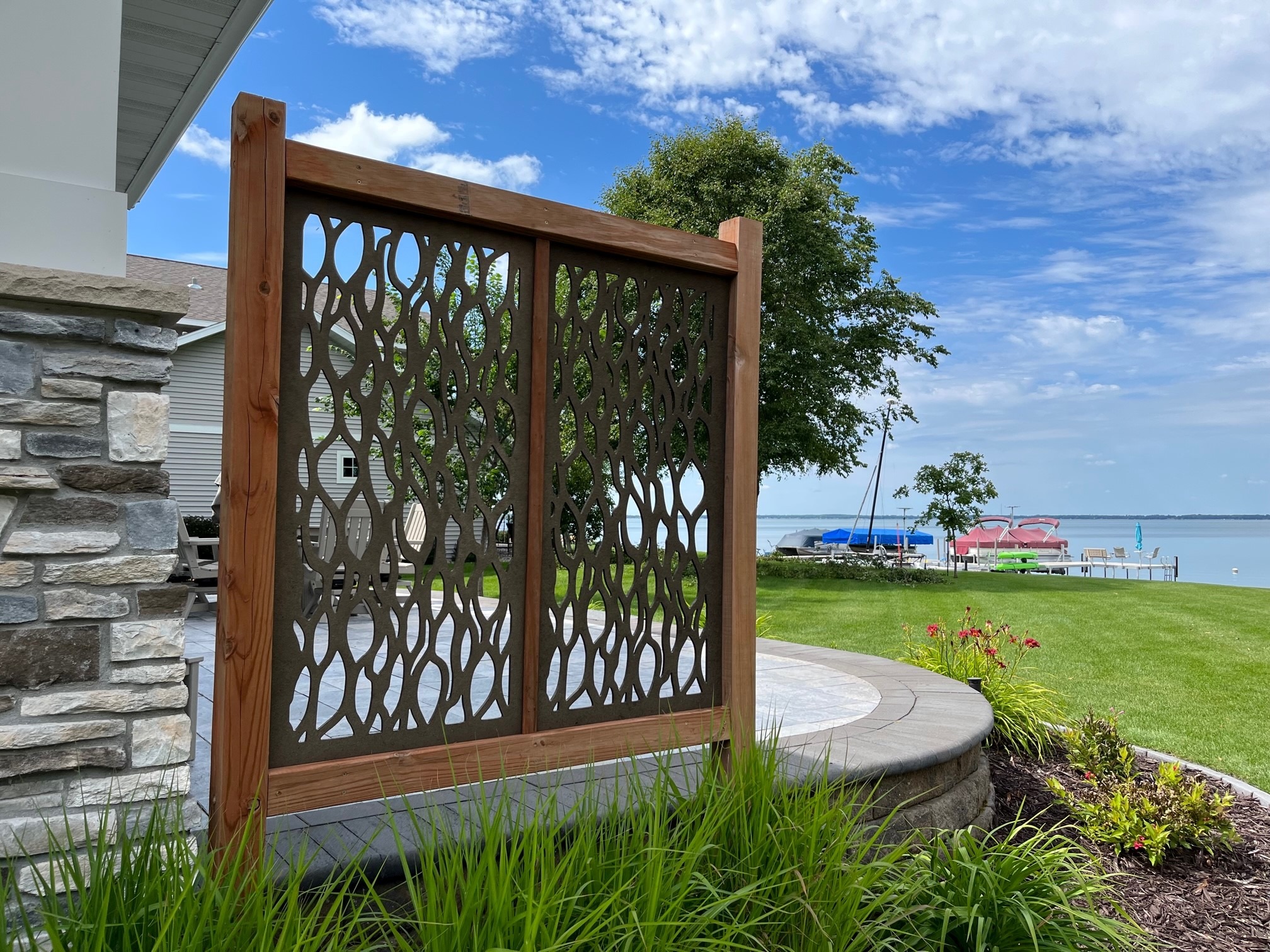 A decorative wooden screen and patio overlook a serene lake, with boats and a dock. Lush greenery and blue sky complete the scene.