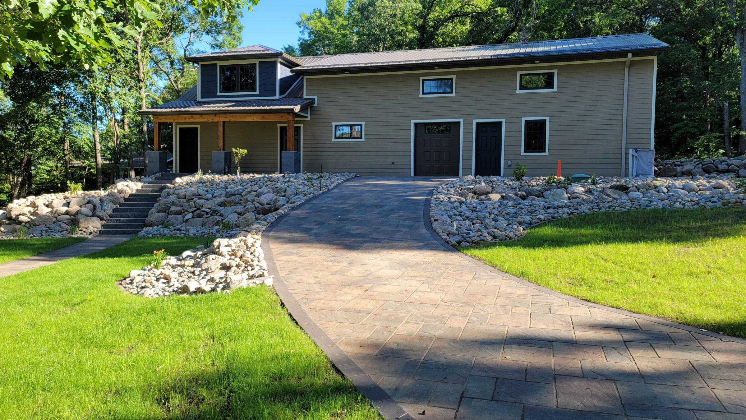Modern house with two stories, surrounded by lush trees, rocks, and neatly trimmed grass. Paved walkway leads to the entrance. Peaceful setting.