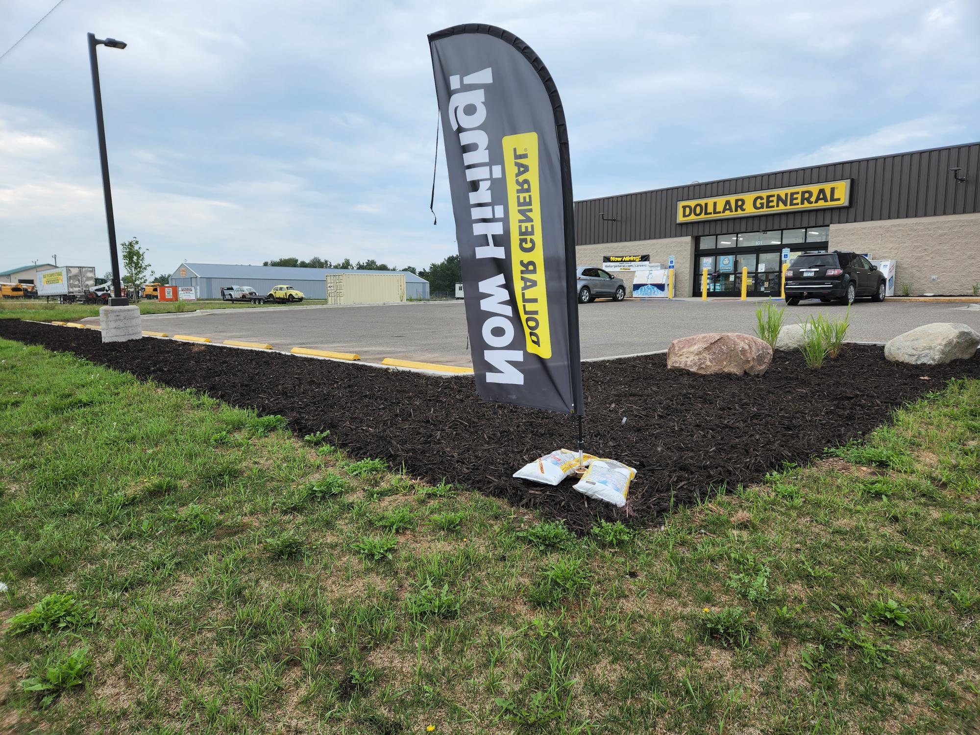 A Dollar General store with a parking lot and a "Now Hiring" flag, accompanied by fresh landscaping and a cloudy sky above.