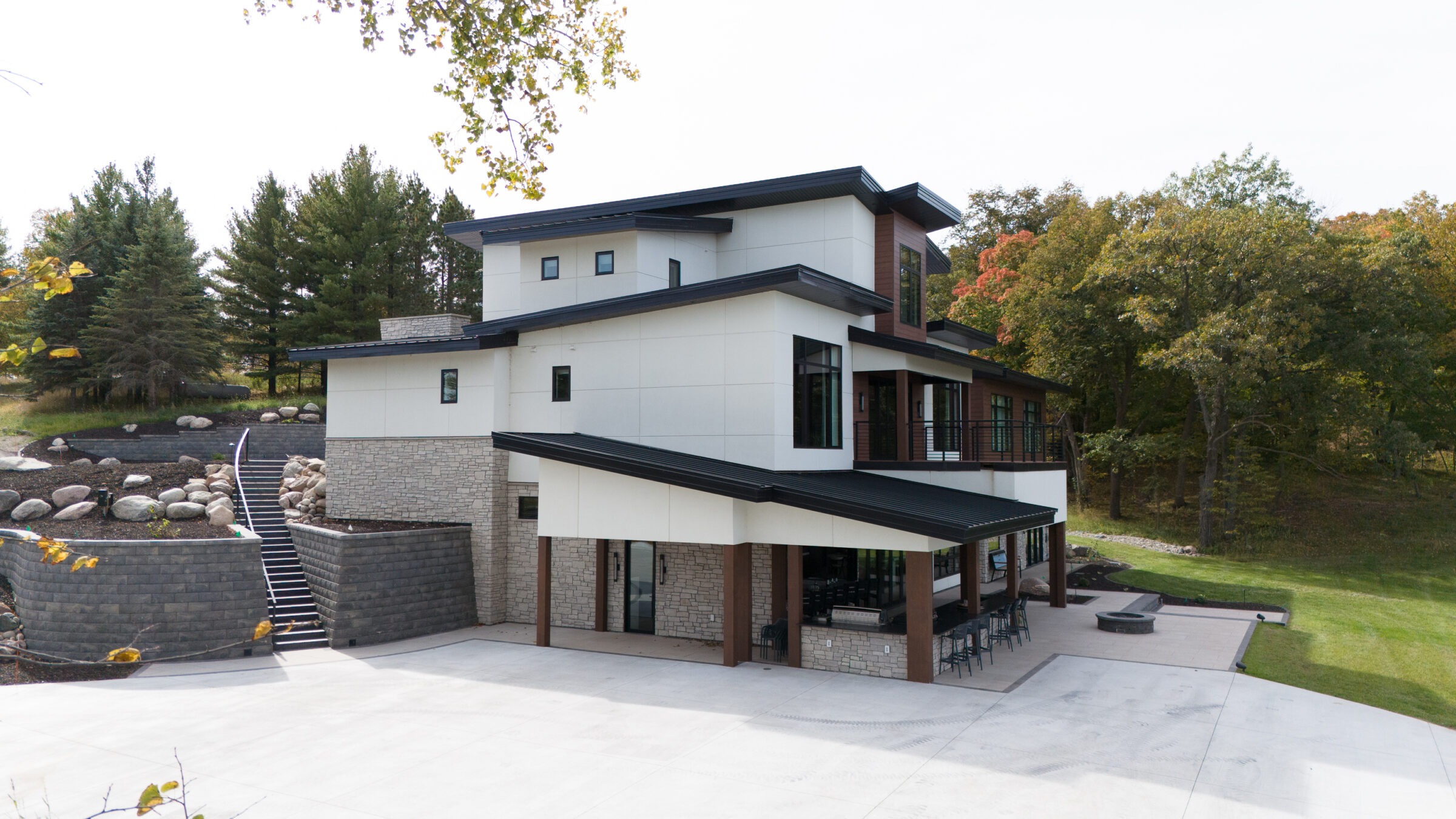 Modern house with stone and wood elements, surrounded by trees. Outdoor staircase leads to landscaped area with large rocks. Spacious patio visible.