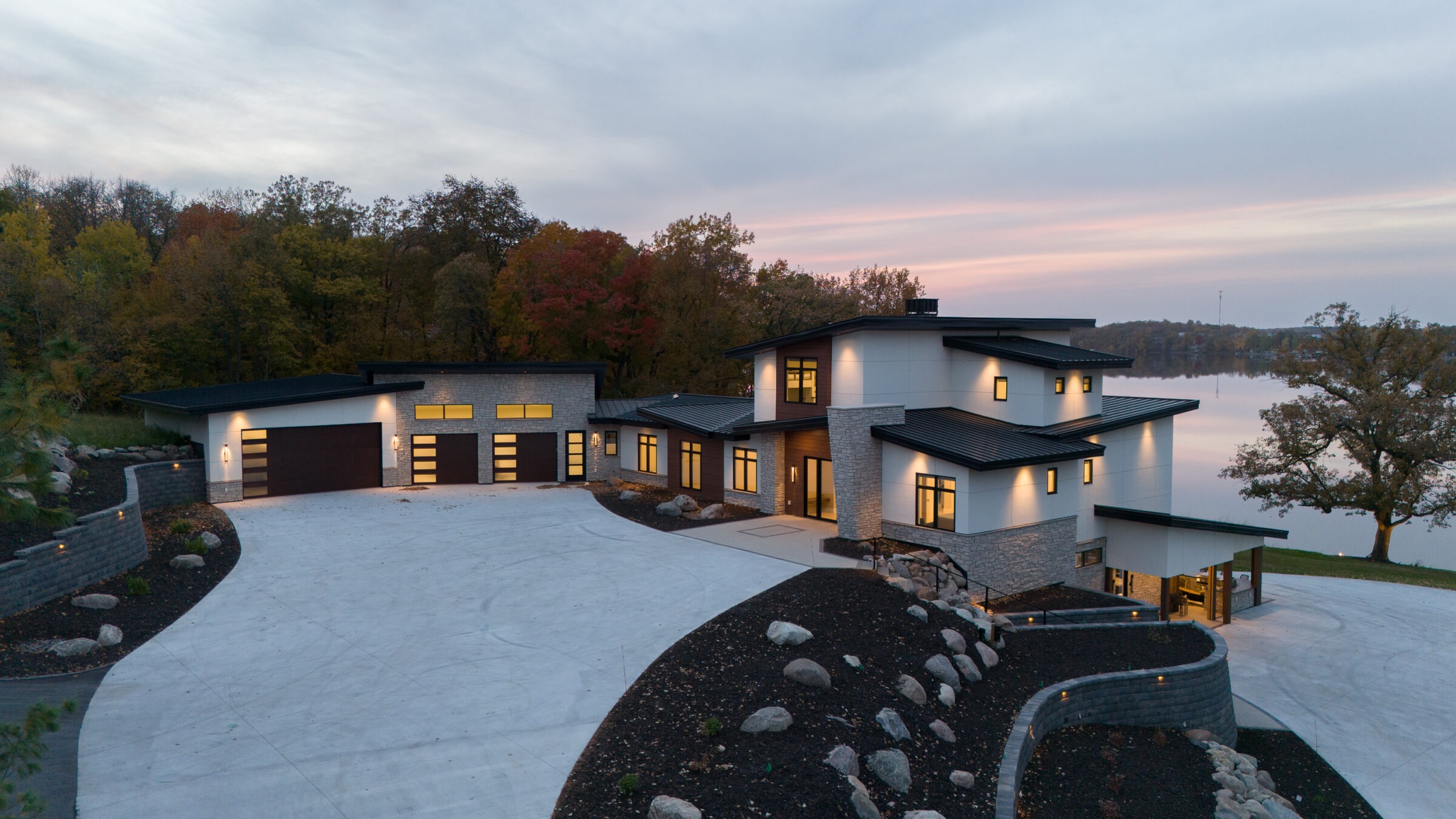 Modern lakeside house with large windows, surrounded by autumn trees and a spacious driveway, reflecting warm interior lighting at dusk.