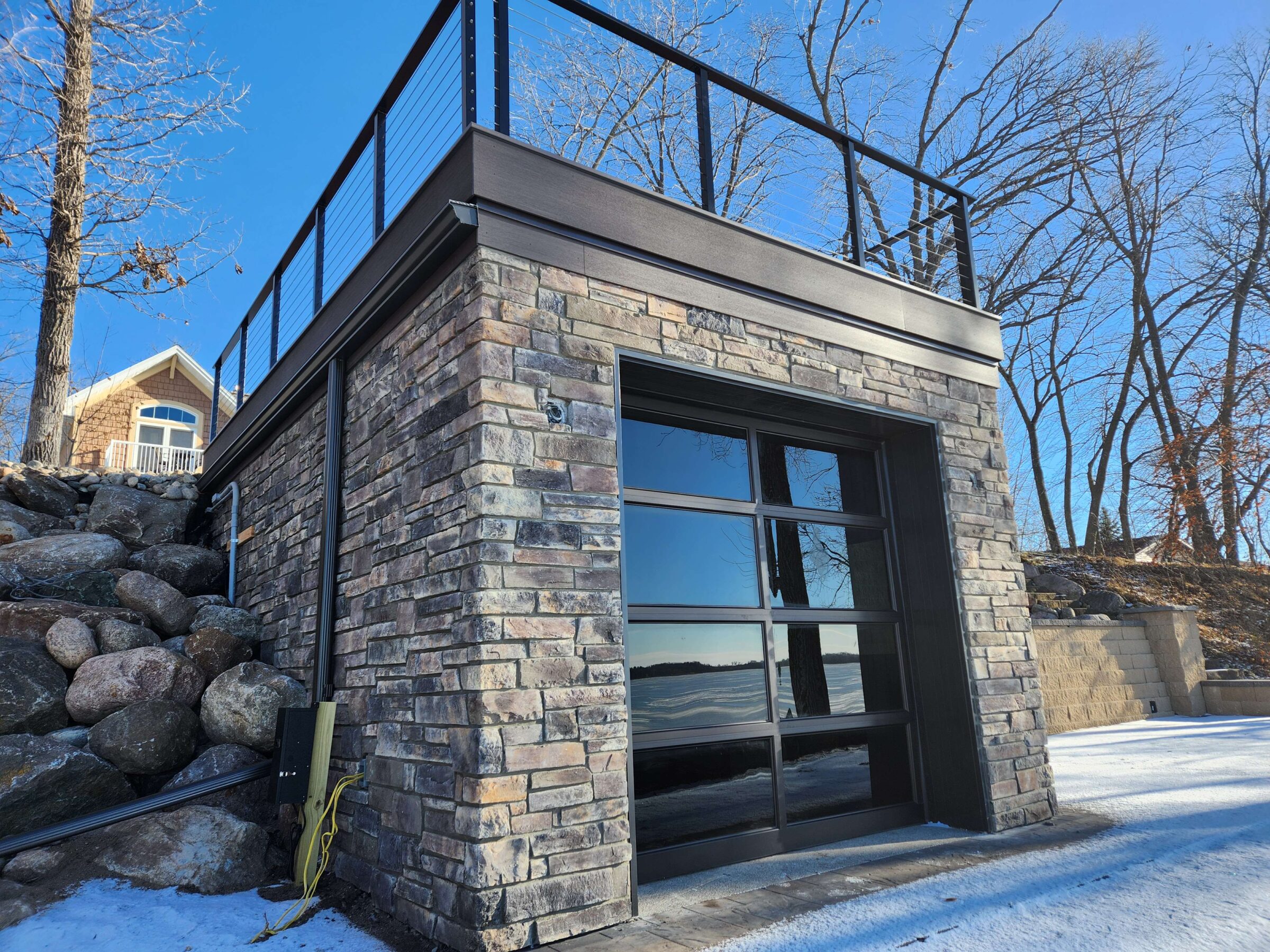 A stone garage with large windows reflects a snowy, wooded landscape. Nearby, a house is visible under a clear blue sky.