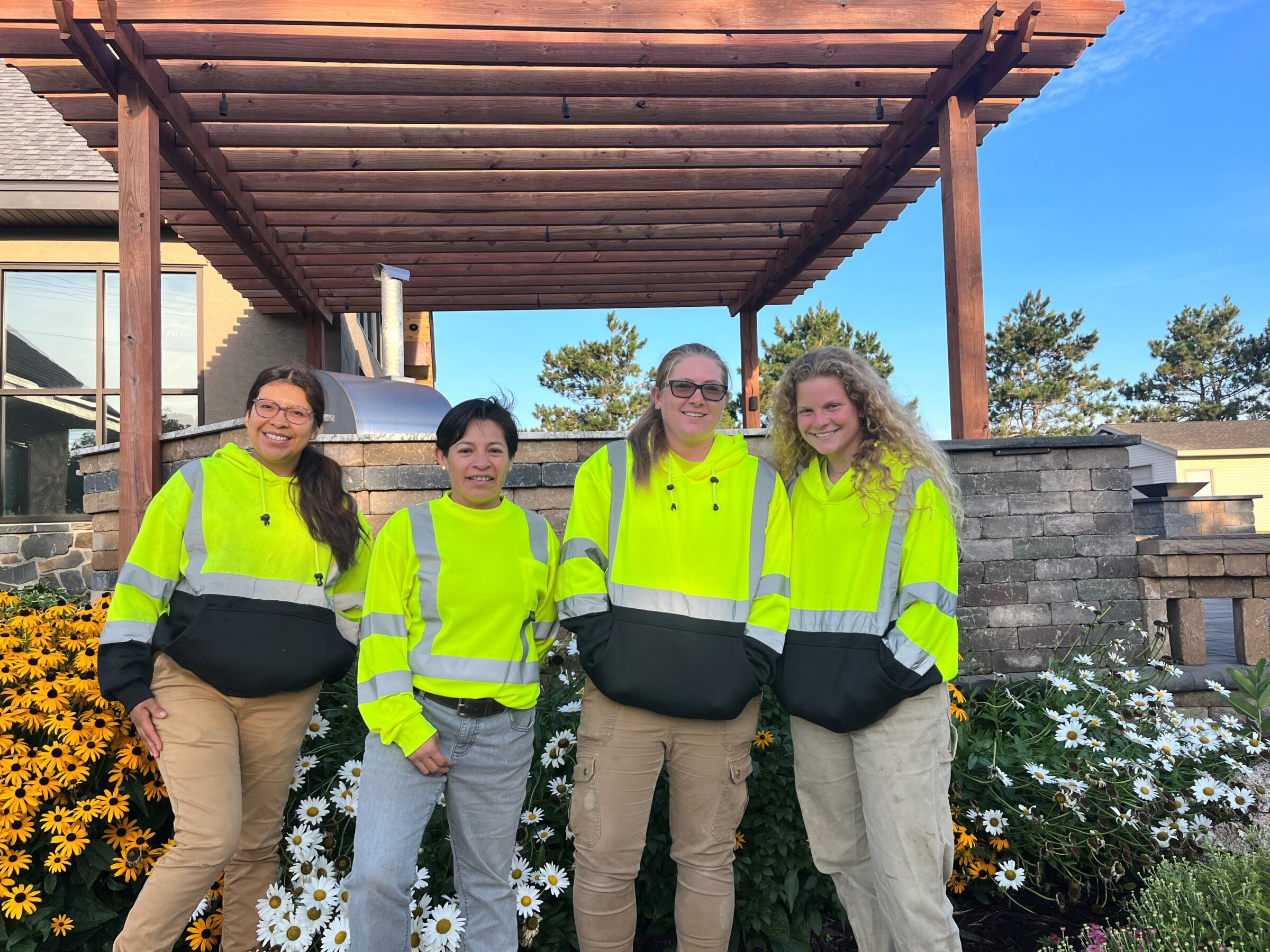 Four people in bright reflective jackets stand together in front of a flower garden and wooden pergola on a sunny day.