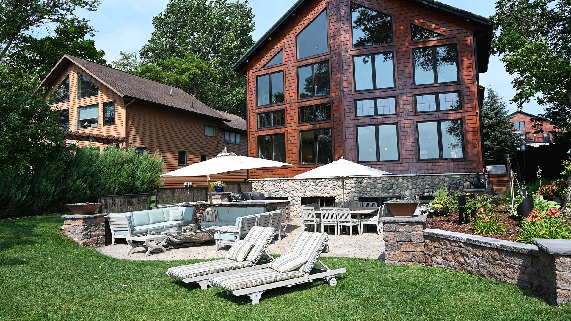 Spacious backyard with loungers, seating area, and umbrellas. Two large wooden homes surrounded by greenery and neatly arranged gardens on a sunny day.
