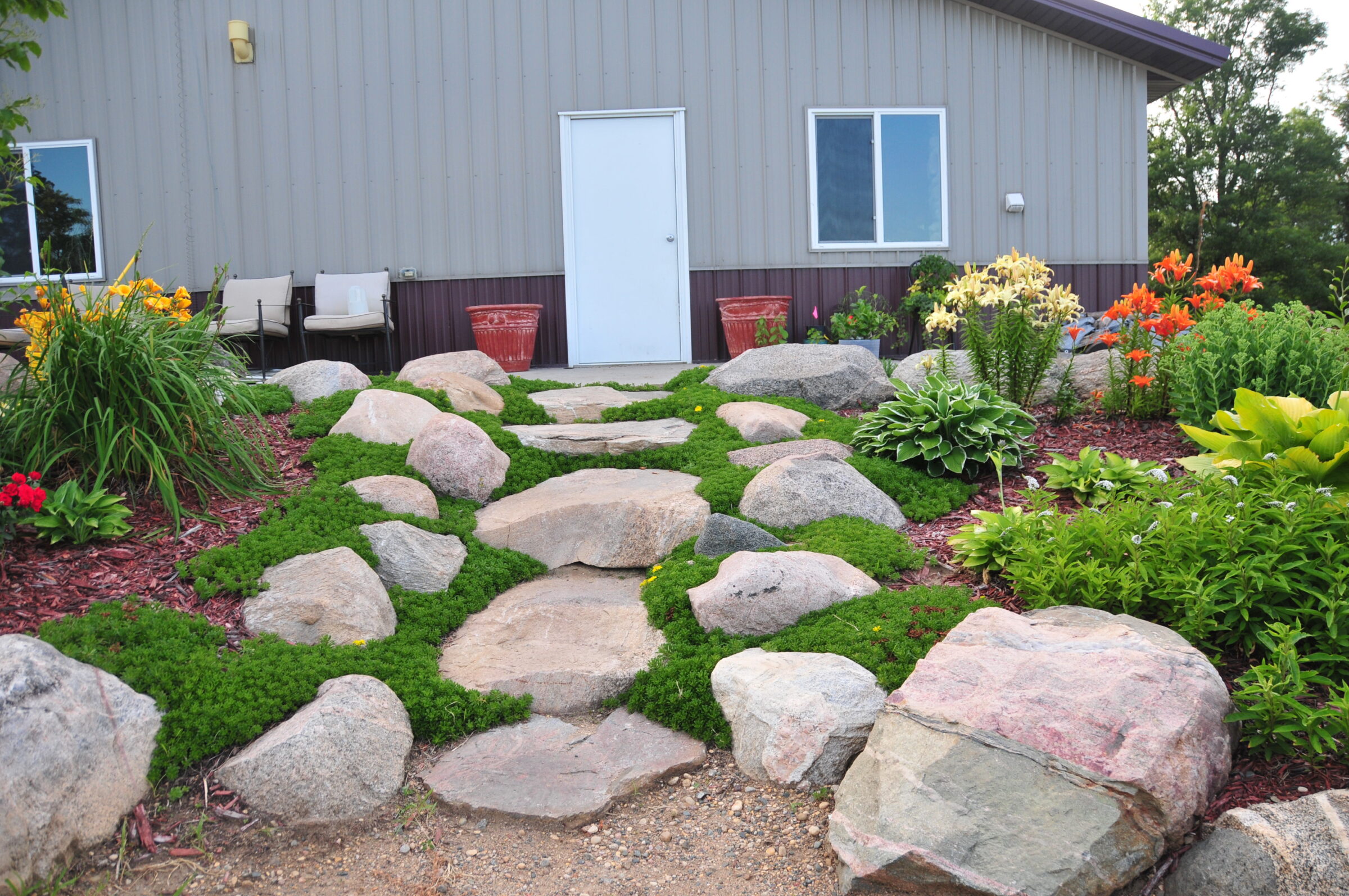 A garden path with rocks and colorful flowers leads to a door of a grey building, surrounded by lush greenery.