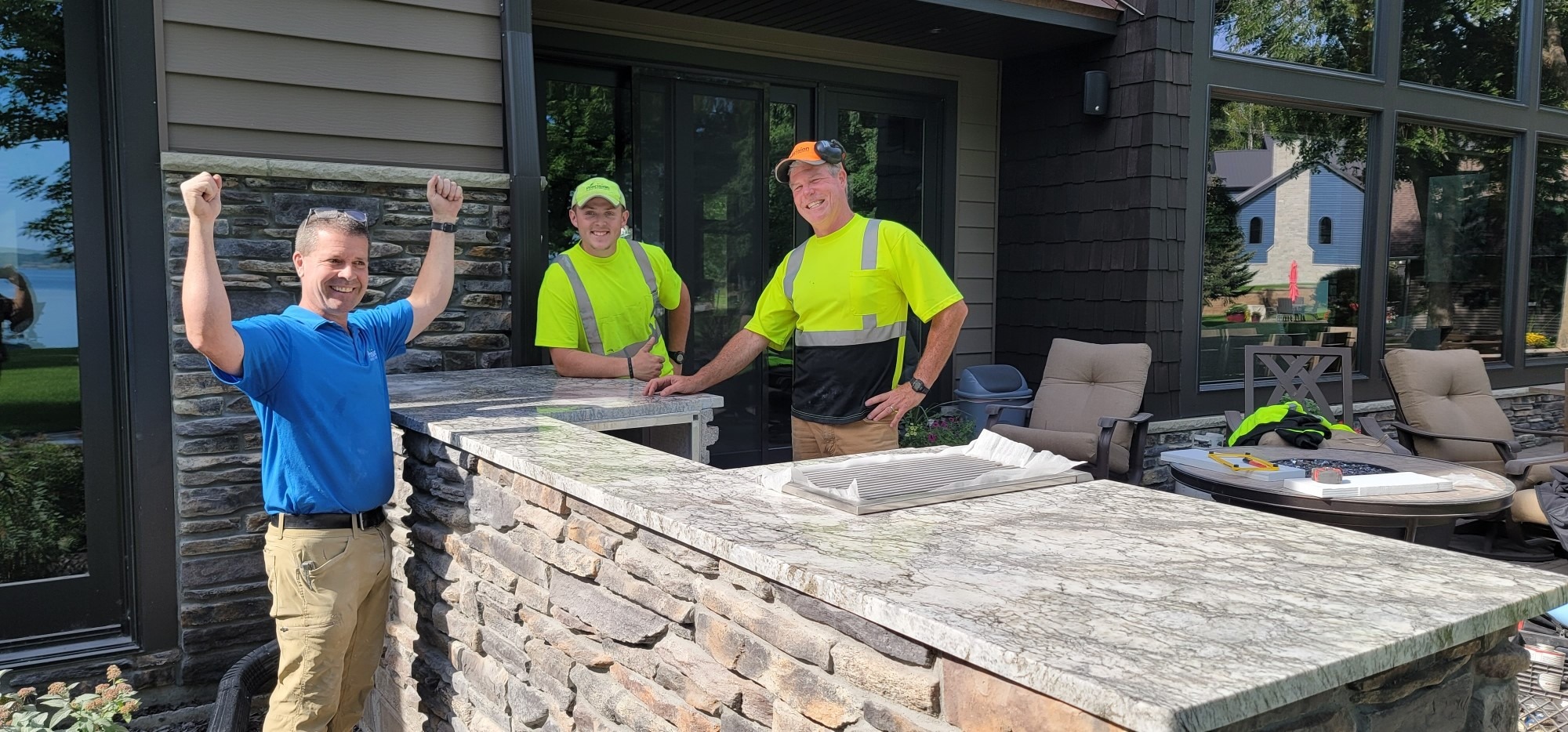 Three people standing outside by a new stone counter, two in work attire, one celebrating. A house and lawn visible in the background.