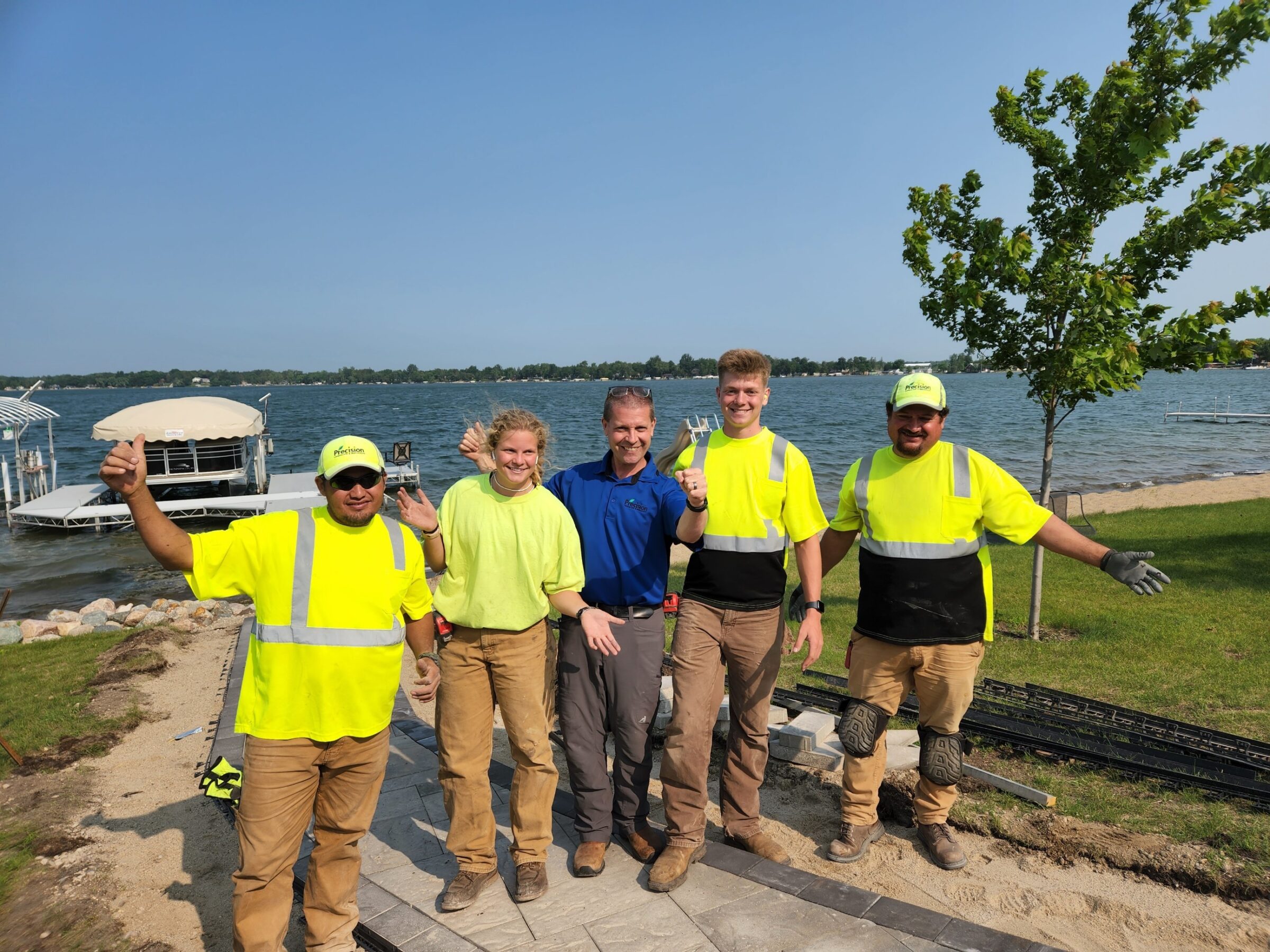 Five people in bright work shirts pose happily by a lakeside, with a docked boat and calm water in the background.