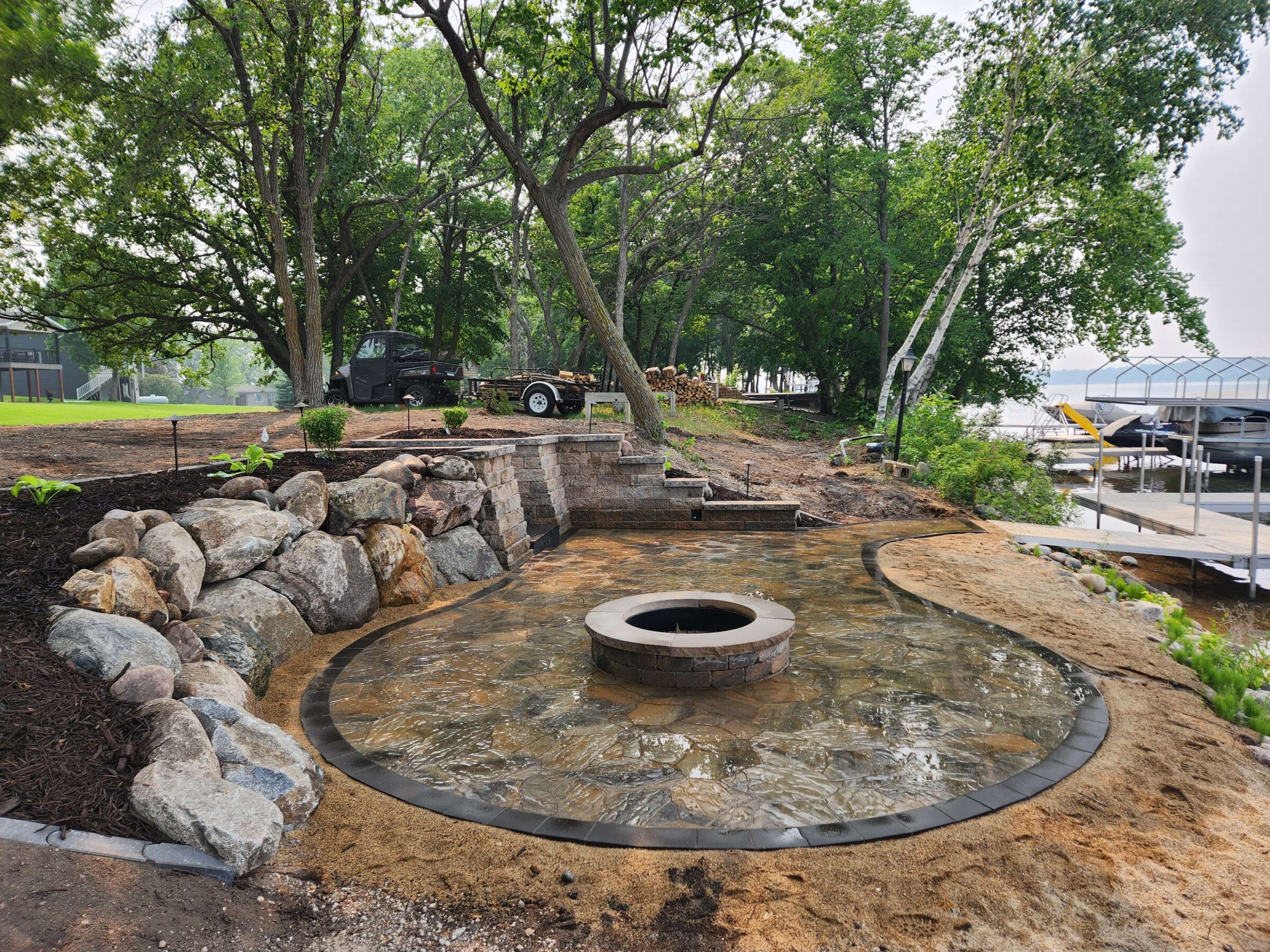 A serene lakeside scene with a stone fire pit, surrounded by trees, rocks, and docks, under a clear sky.