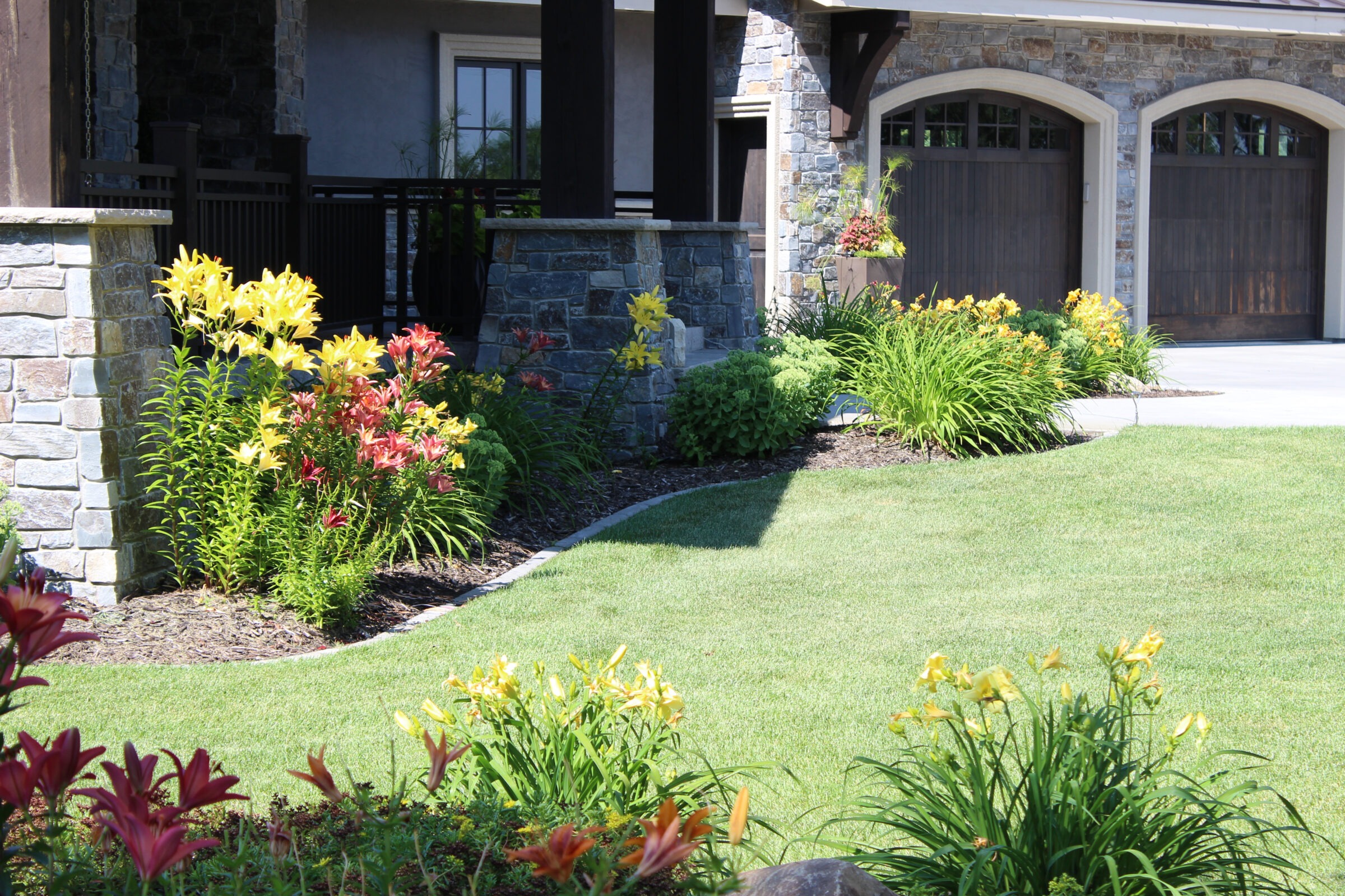 A stone house with a manicured lawn is surrounded by colorful flowers and shrubs, leading to a wooden garage door.