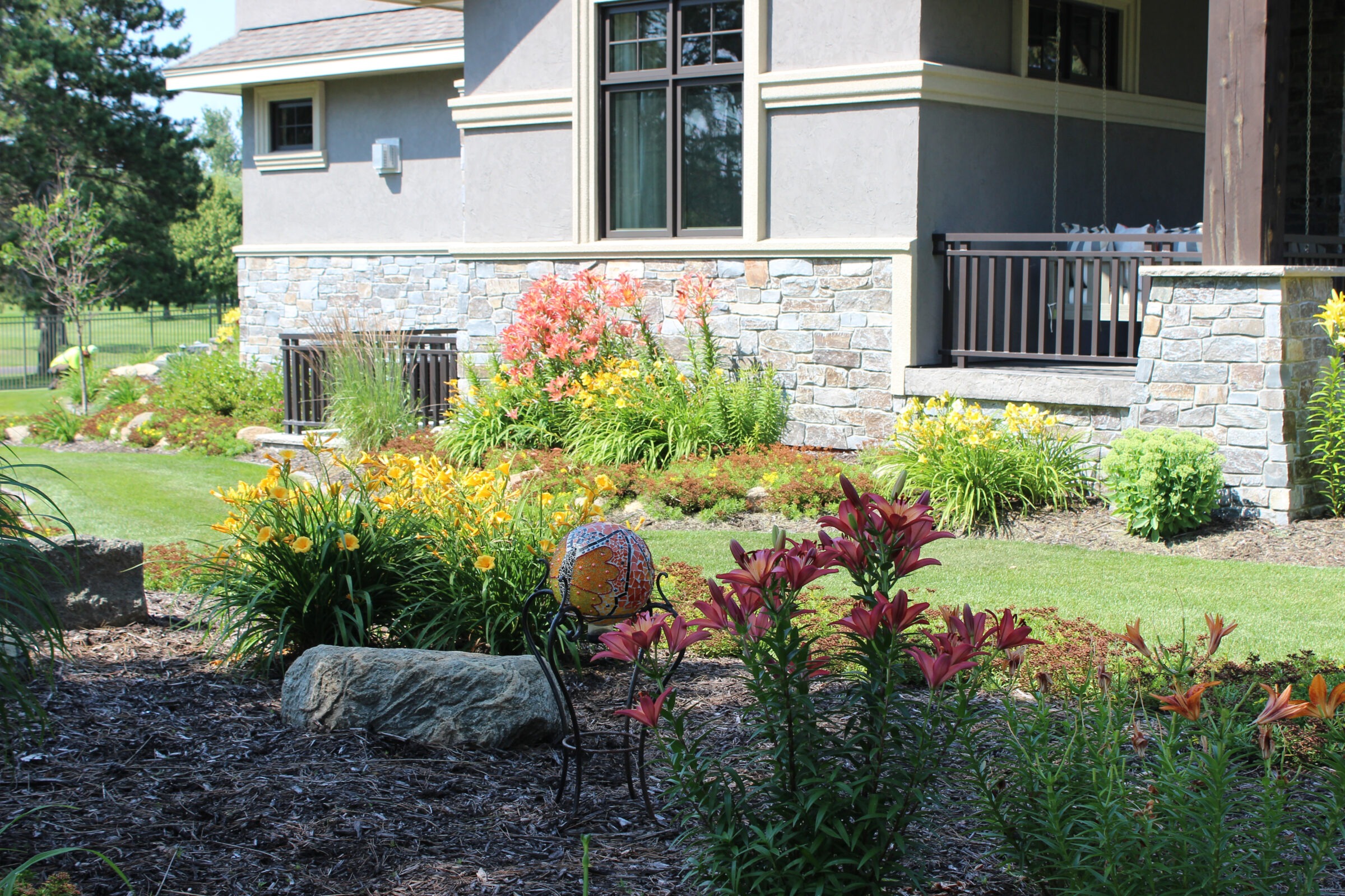 A garden with colorful flowers and stone accents surrounds a building with a patio. A person tends to the landscaping.