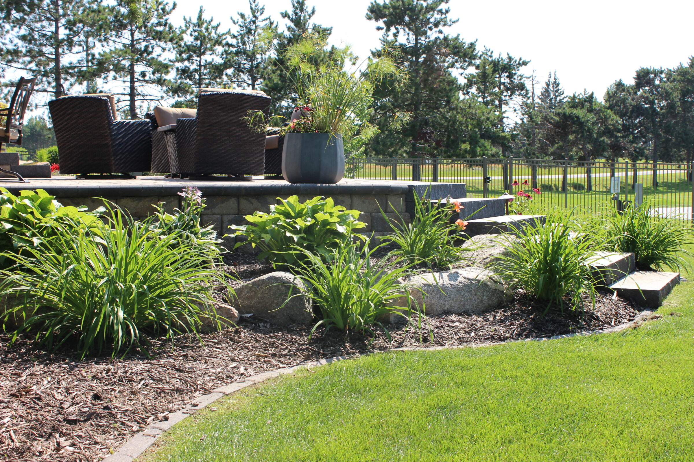 A cozy patio area with wicker chairs overlooks a landscaped garden with lush green plants and stone steps, surrounded by a wooden fence.