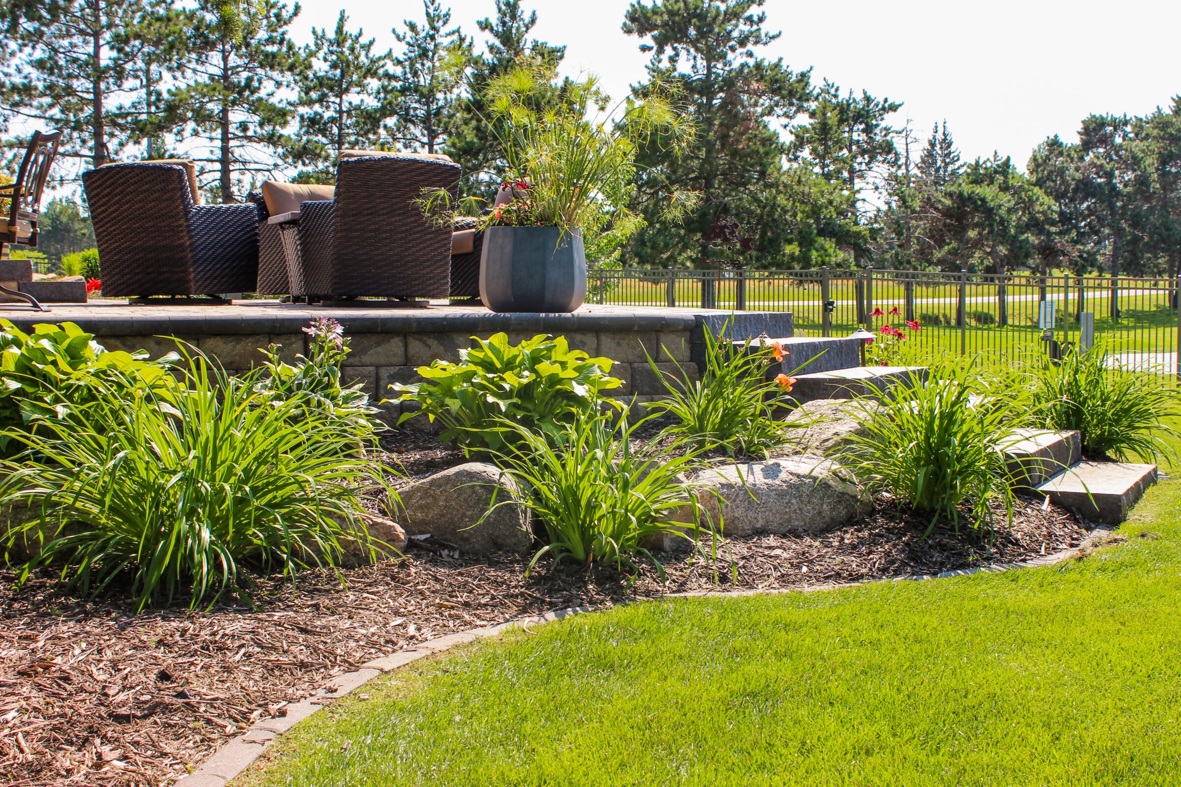 A landscaped garden with lush greenery, stone steps, outdoor wicker furniture, and a potted plant under a clear sky surrounded by trees and a fence.
