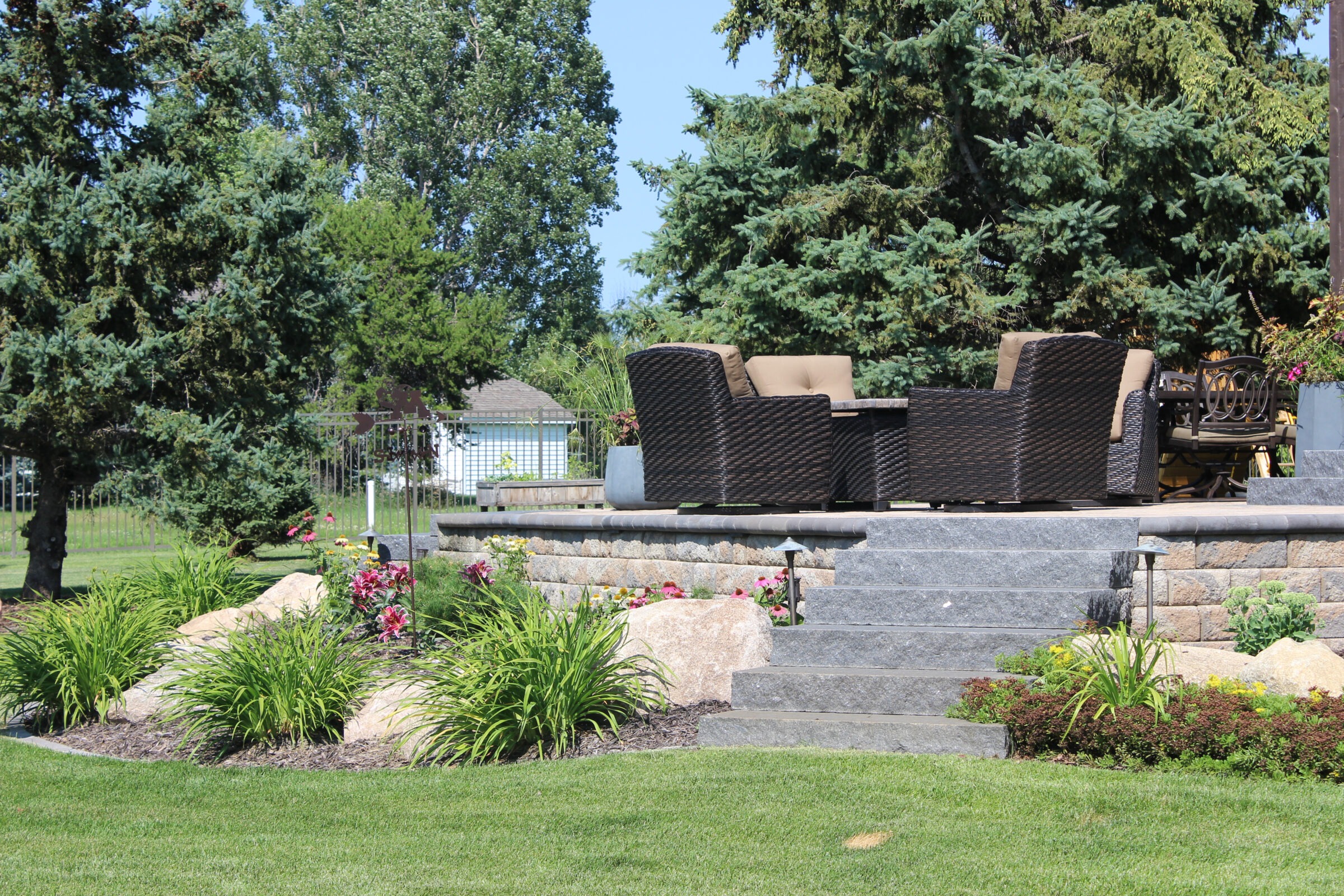 Garden with stone steps leads to outdoor seating area, surrounded by lush greenery and trees under a clear blue sky.