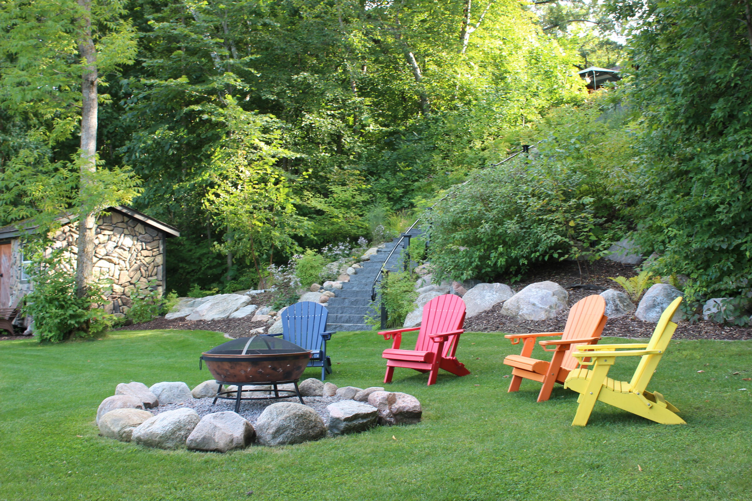 Colorful Adirondack chairs encircle a stone-lined fire pit in lush garden setting, near stone shed and stairway leading into dense greenery.