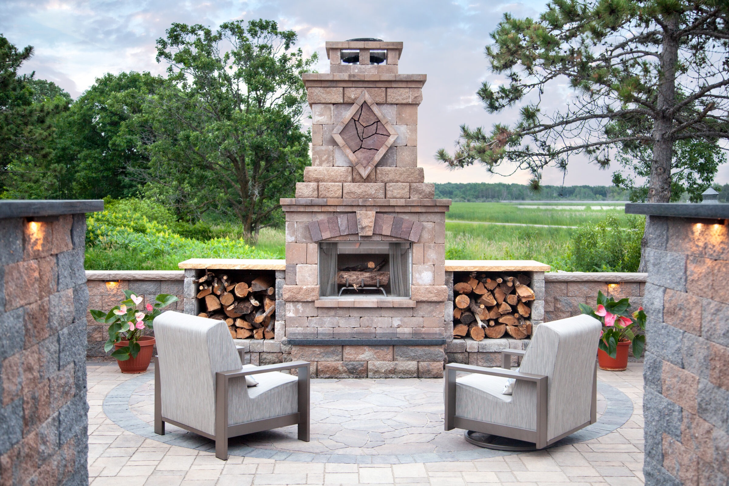 Two chairs face an outdoor stone fireplace, surrounded by greenery. Firewood and potted plants complete the serene patio setting.