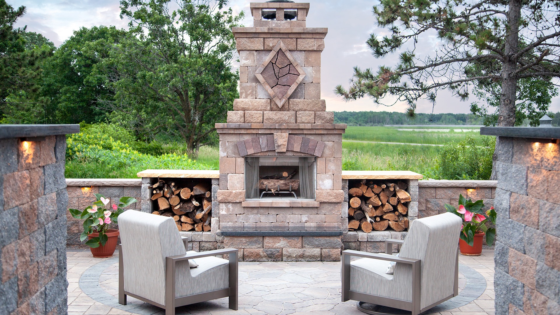 Outdoor patio with stone fireplace, two chairs, stacked firewood, and potted plants. Surrounded by lush greenery under a cloudy sky.