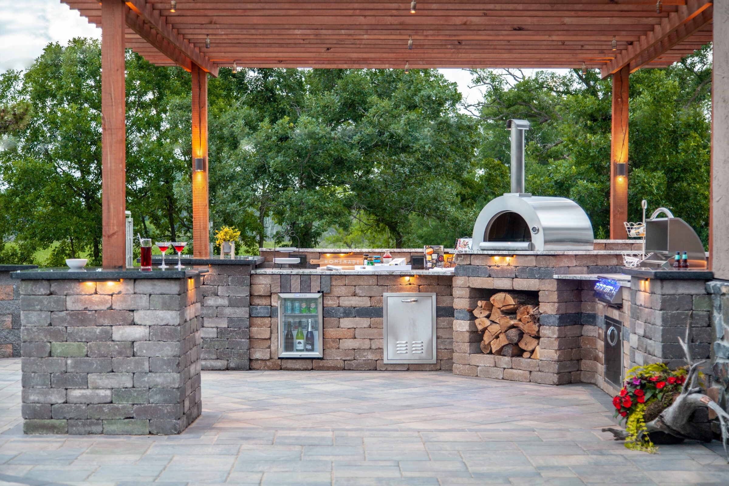 Outdoor kitchen featuring a stone counter, wood-fired oven, grill, and fridge beneath a wooden pergola, surrounded by lush trees.