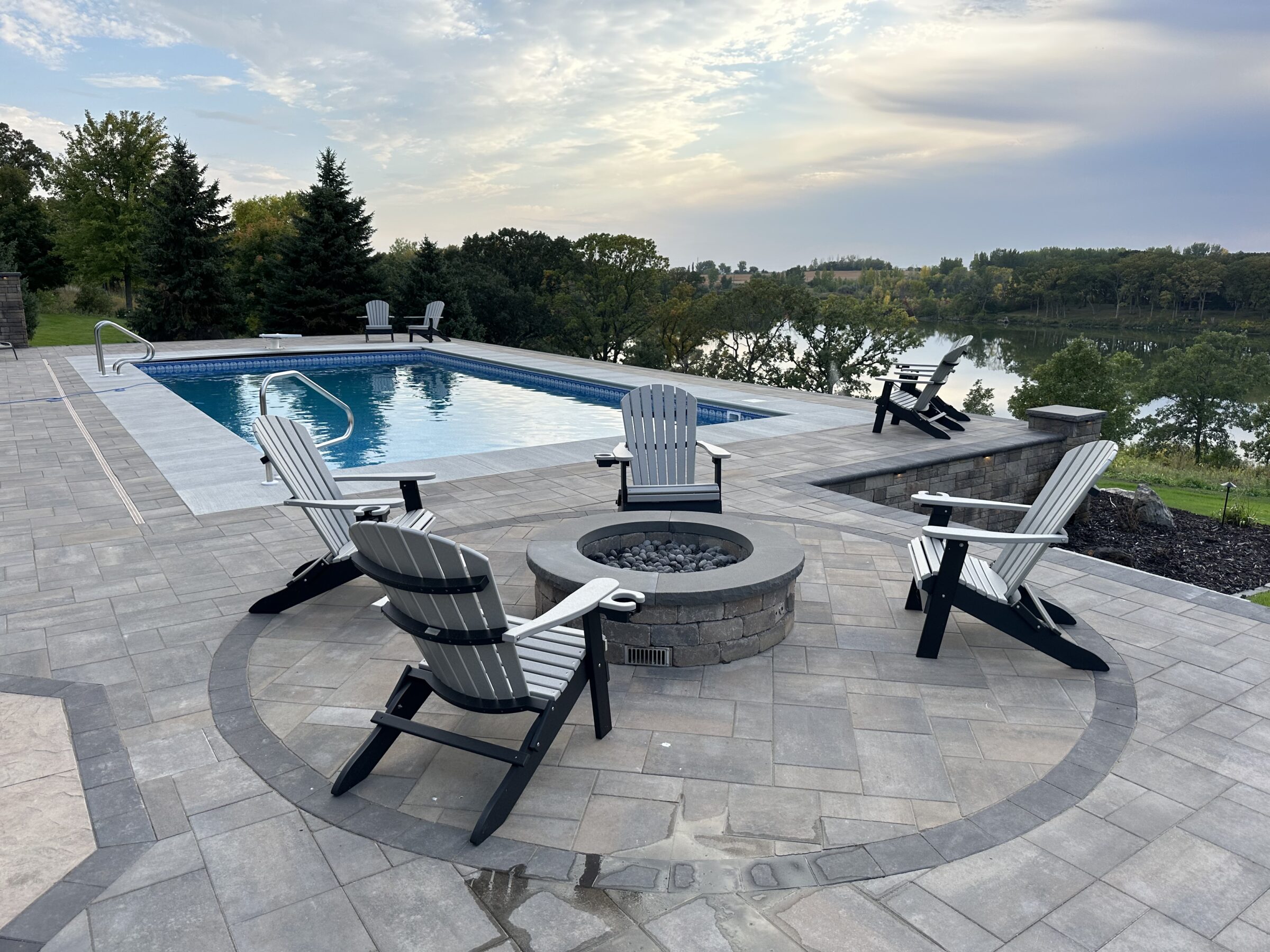 A serene patio with a pool, surrounded by Adirondack chairs and a fire pit, overlooking a calm lake and lush trees.