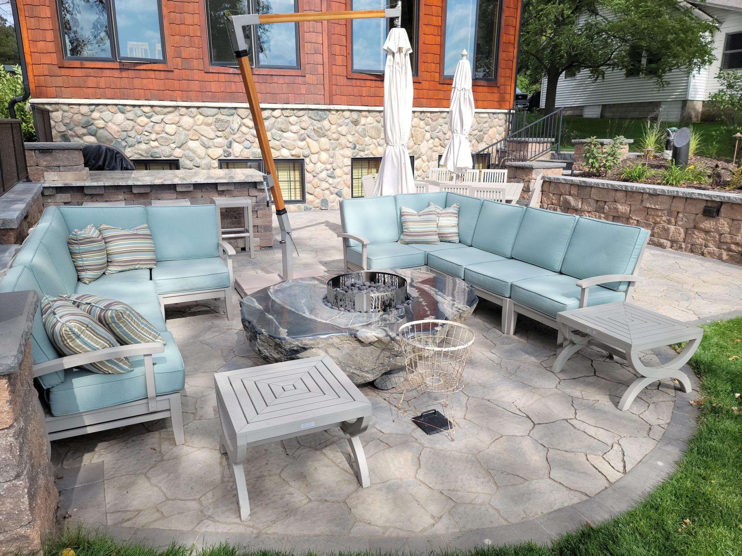 Outdoor patio with stone fireplace, blue cushioned seating, and striped pillows. Stone and wood house facade in the background. Peaceful garden setting.