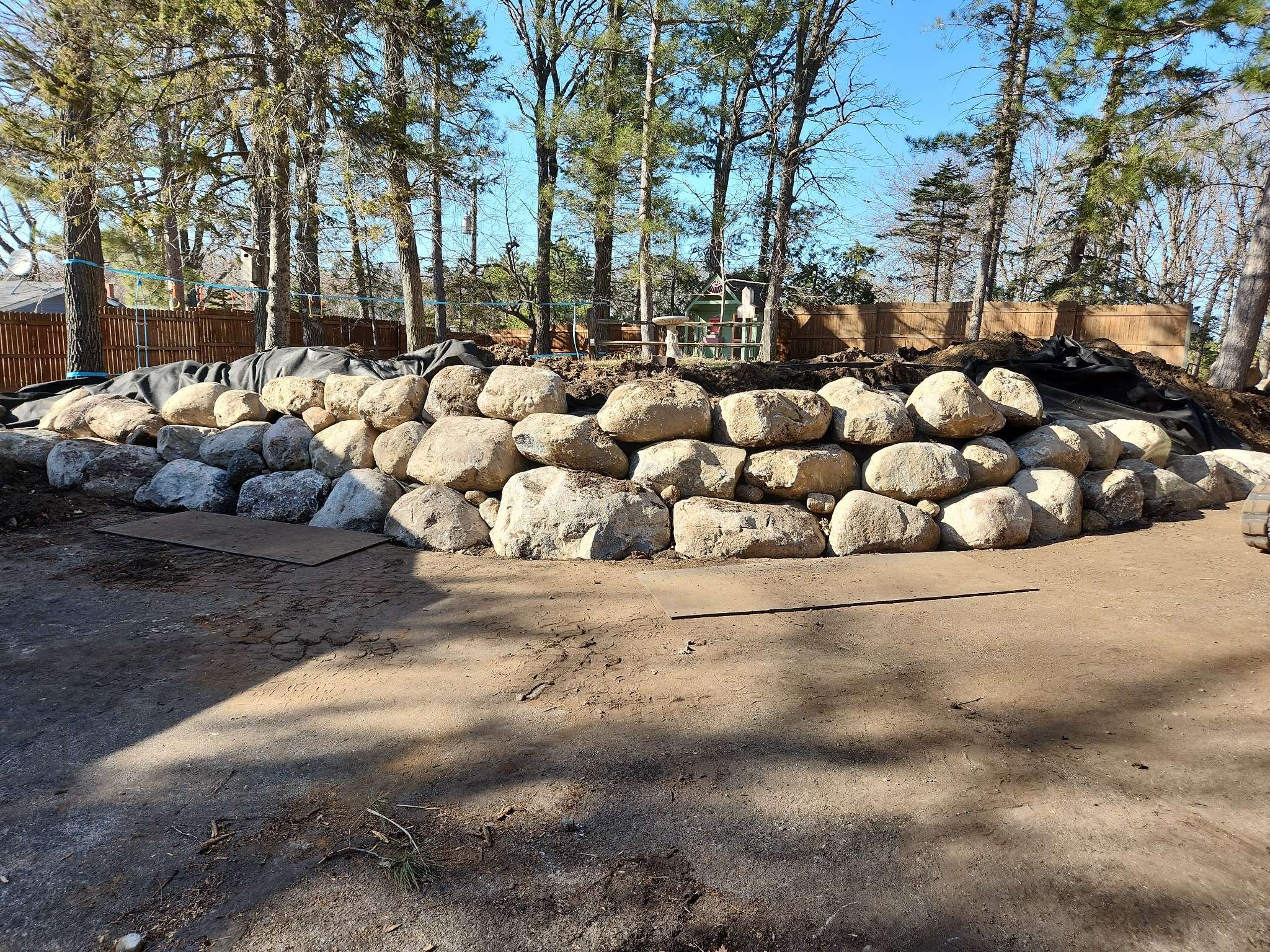 A pile of large rocks forms a retaining wall among trees. A wooden fence and small playground structure are visible in the background.