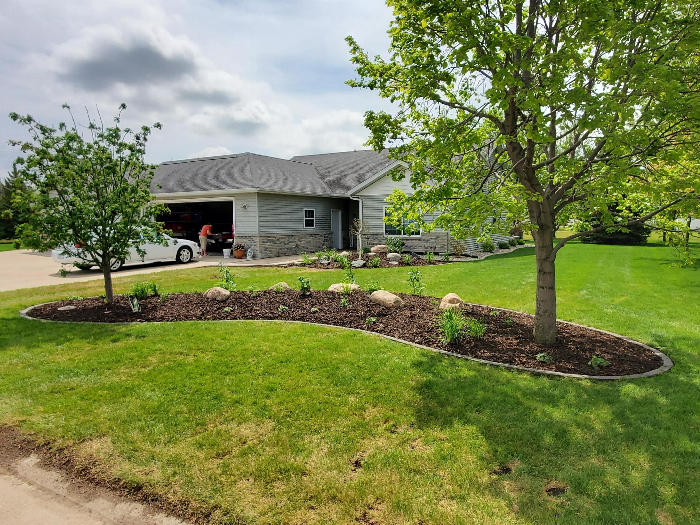 A person stands beside a white car near a suburban house. The lawn is landscaped with trees and a curved flower bed.