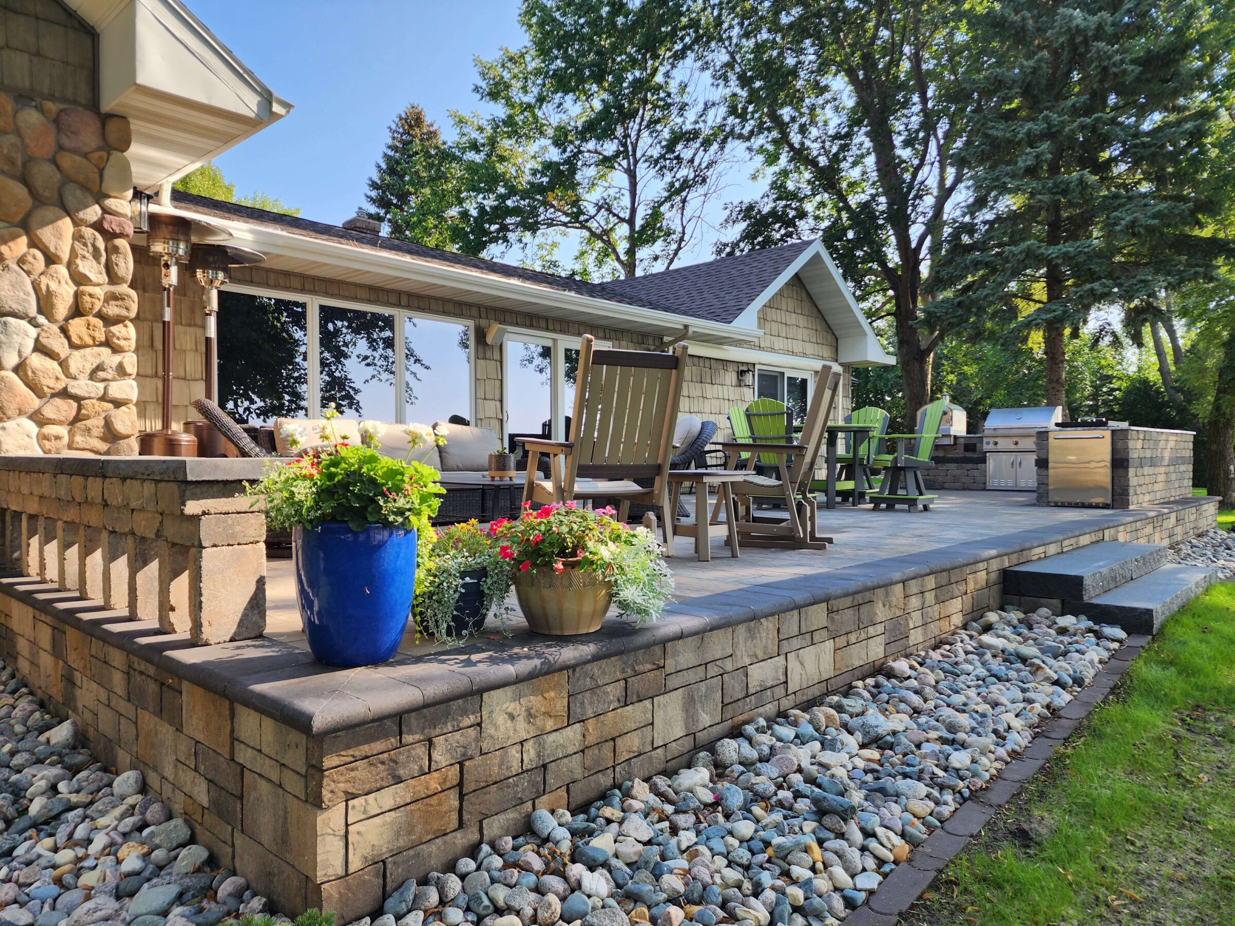 A cozy outdoor patio features a stone fireplace, seating, colorful flower pots, a grill, and lush trees under a clear blue sky.