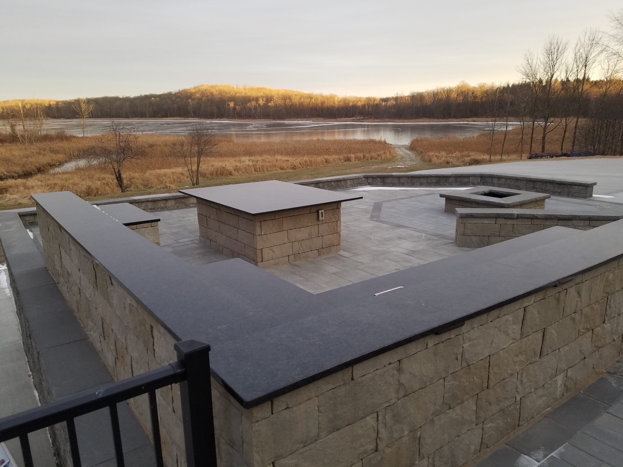 Stone patio with modern sitting area overlooks a serene lake and forested hills under a cloudy sky.