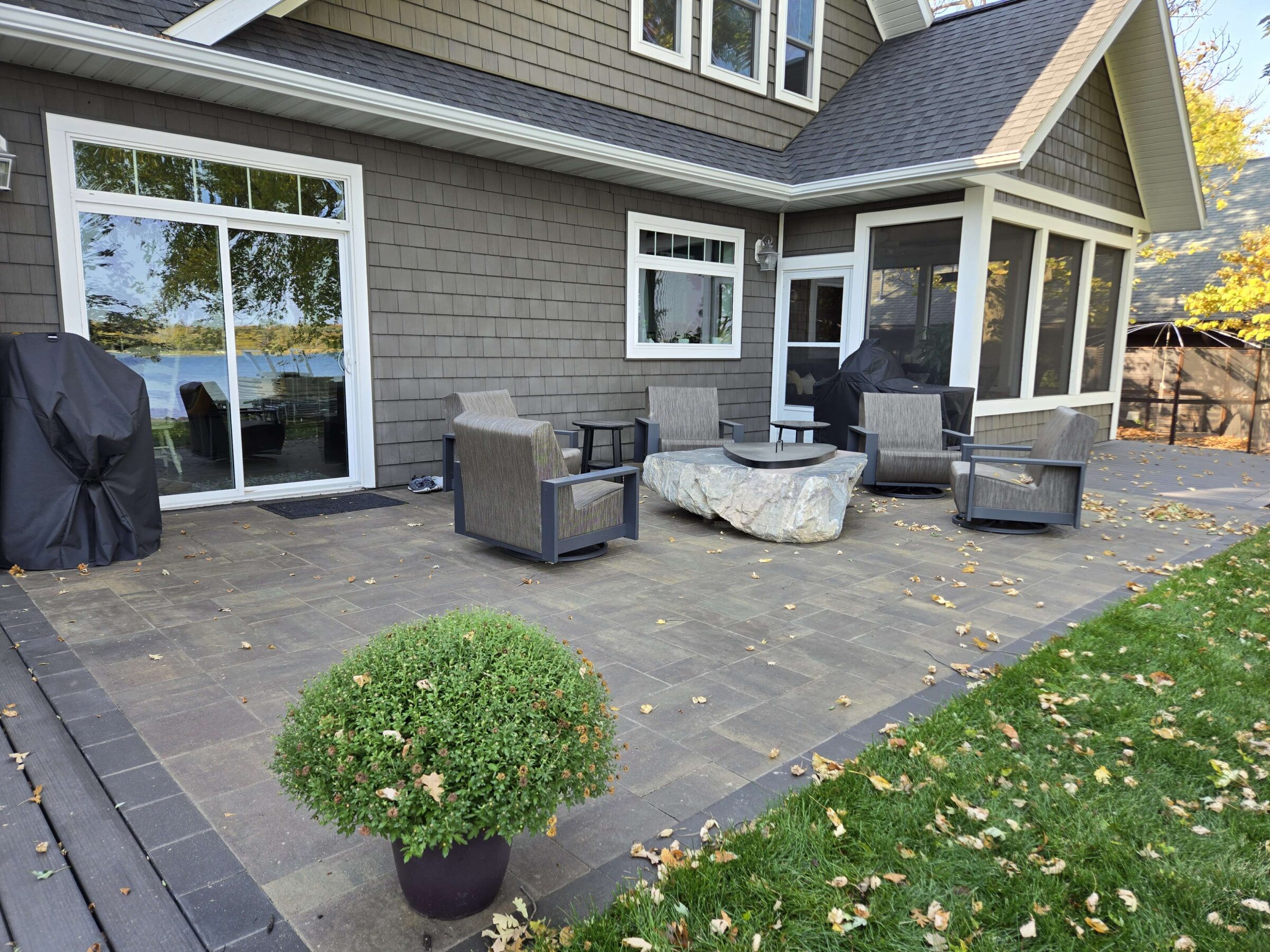 A spacious patio with chairs, a stone table, and a lush plant in front of a brown house overlooking a scenic lake view.