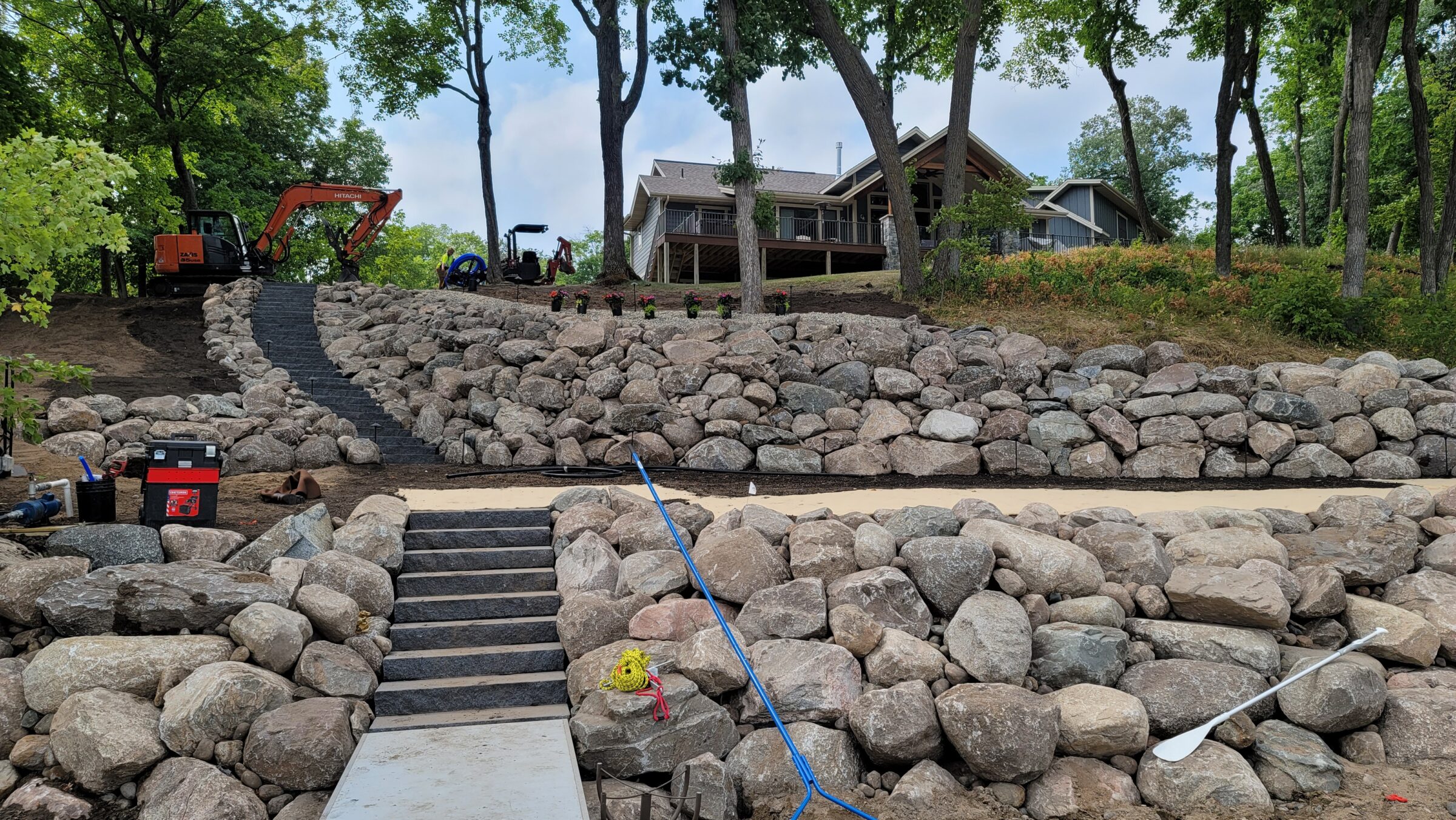 Rock terraces and stairs lead to a house surrounded by trees. Construction equipment and tools are visible, suggesting ongoing landscaping work.