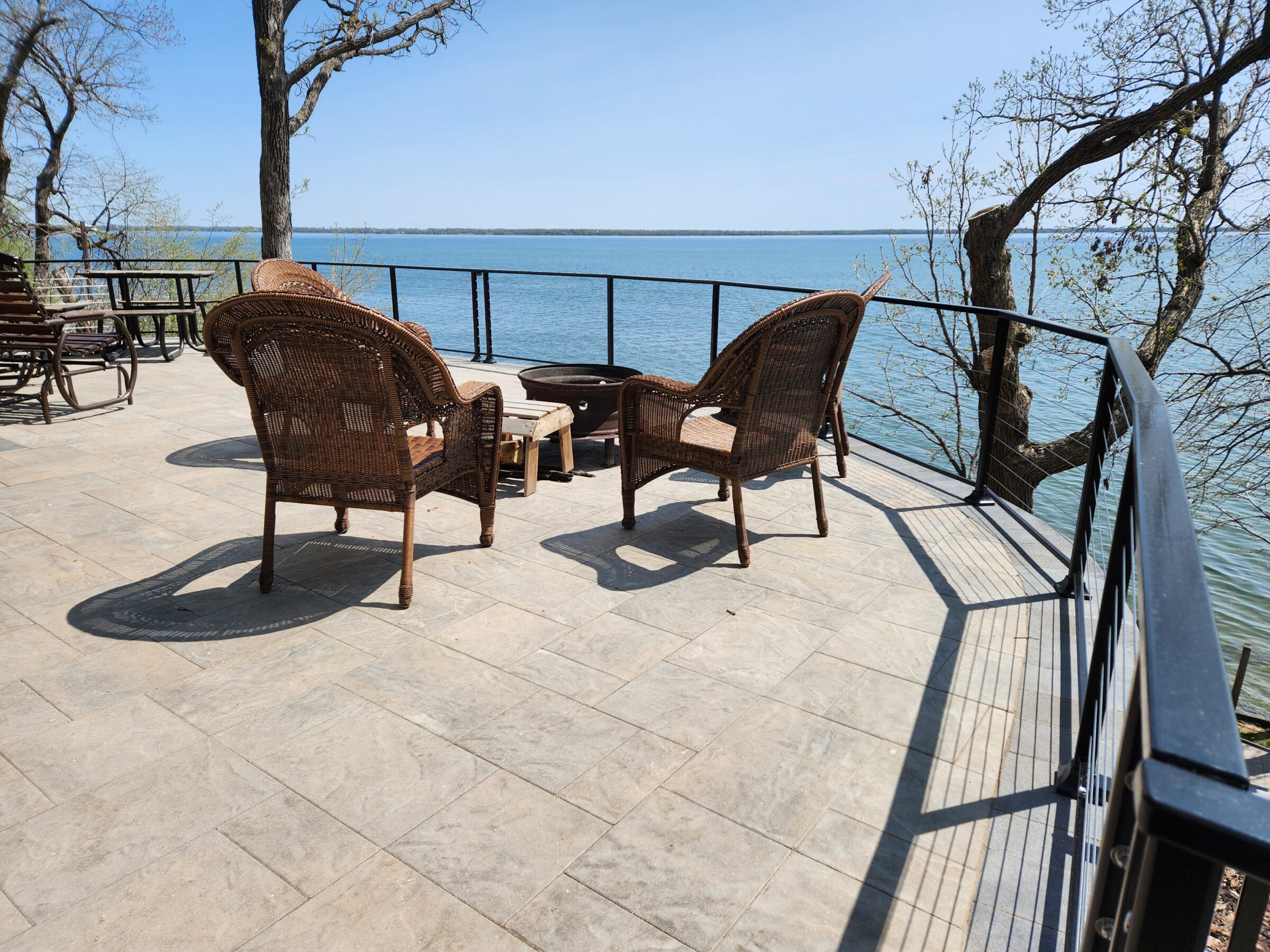 A serene lakeside patio with wicker chairs overlooks a tranquil blue lake, surrounded by trees under a clear sky.