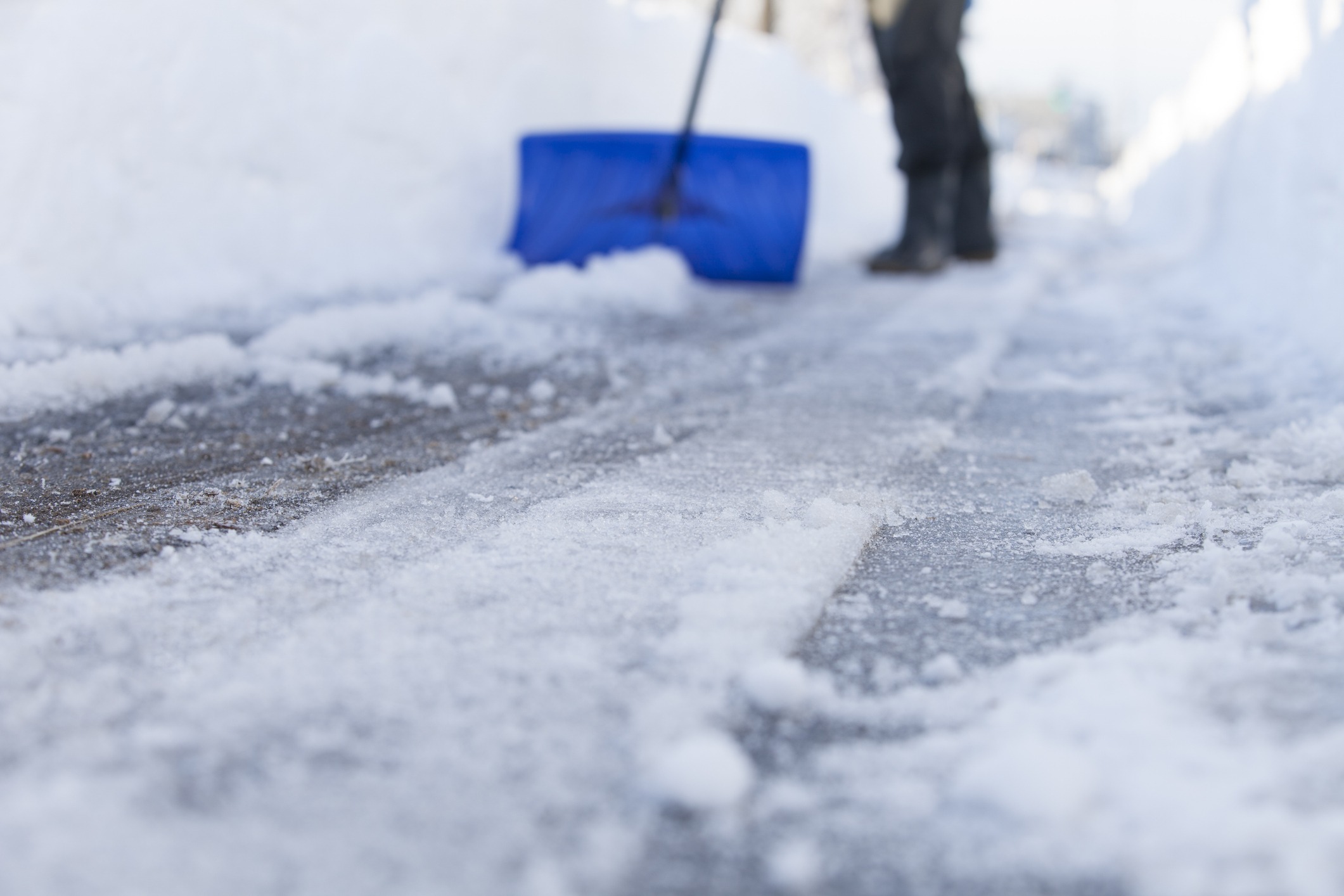 A person shovels snow from a walkway using a blue shovel, creating a clear path surrounded by snowdrifts under a bright sky.