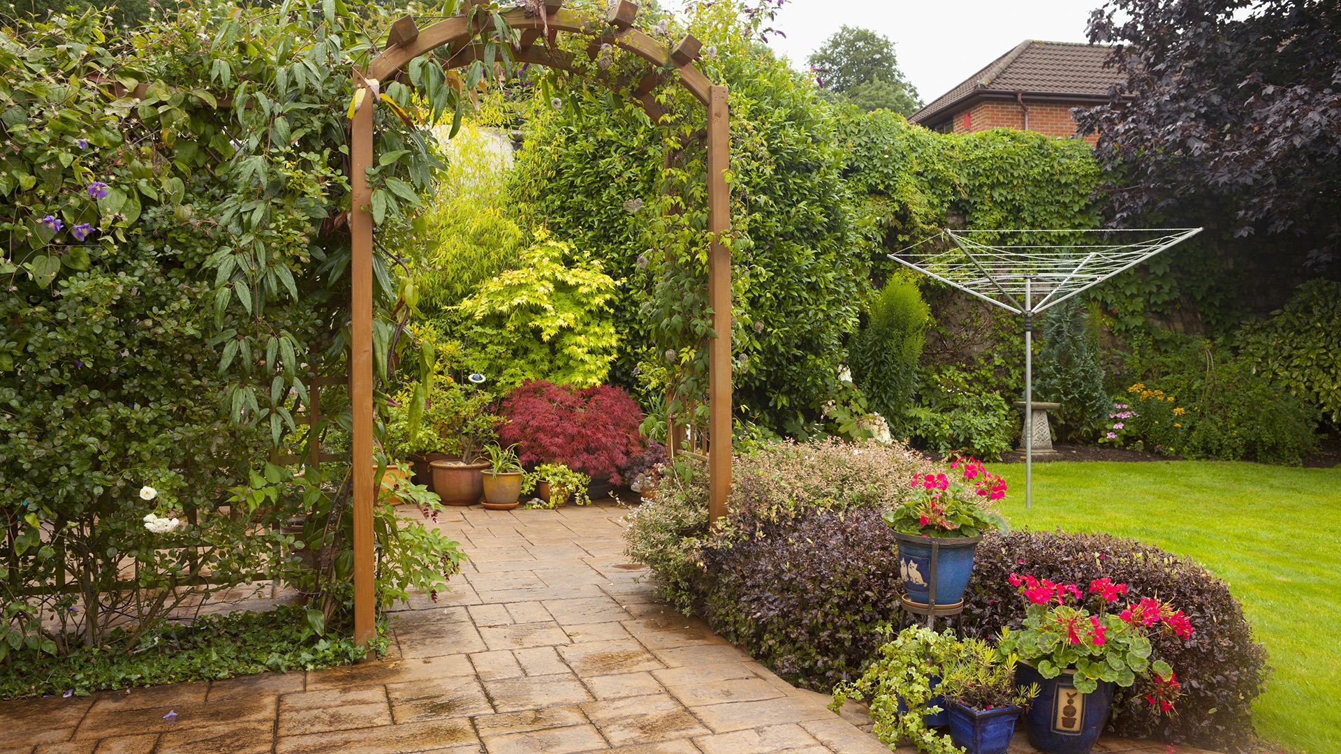 Lush garden with wooden archway, vibrant flowers, and a clothesline. Paved path leads through greenery; vibrant, colorful, peaceful setting.