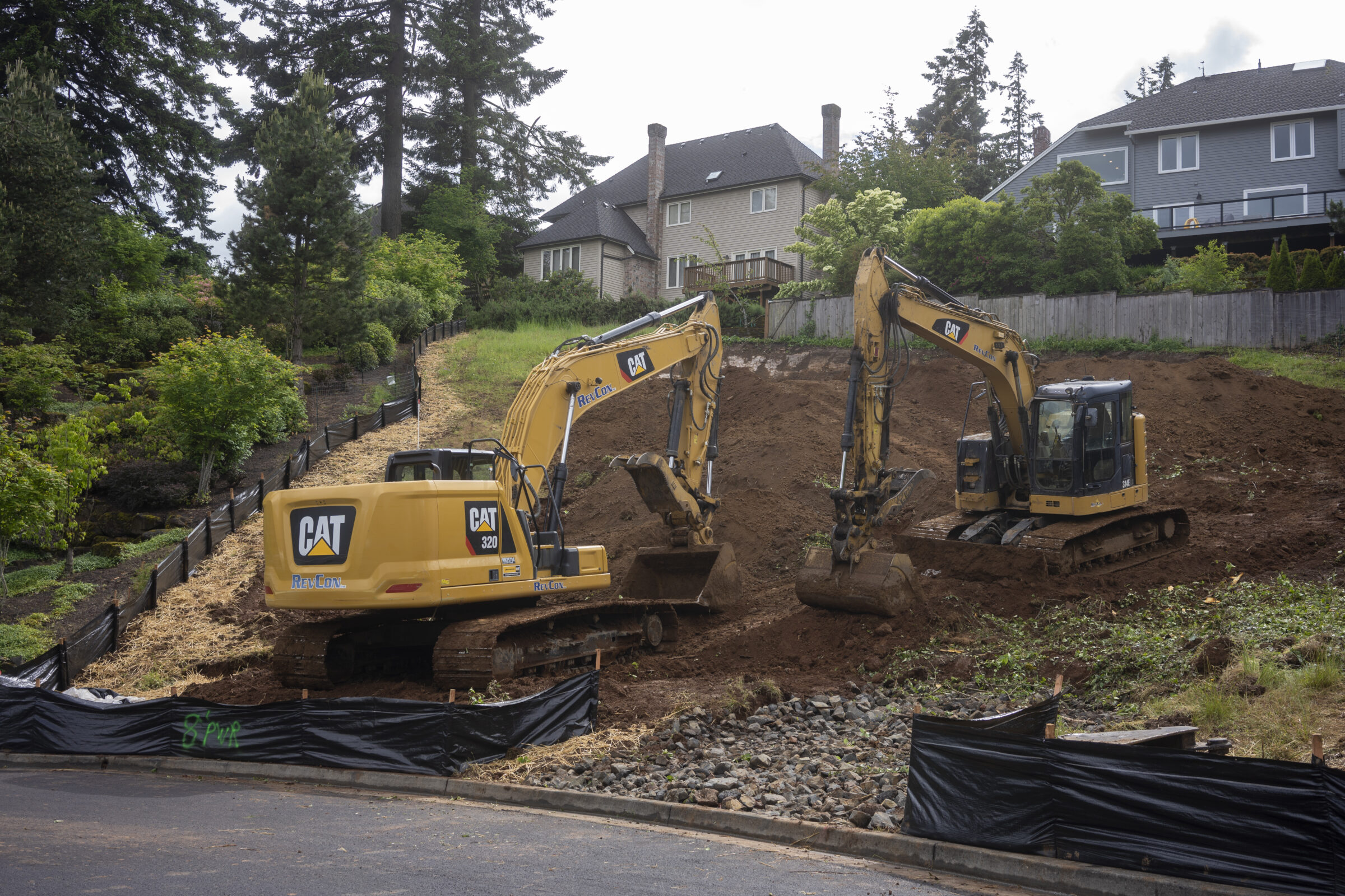 Two excavators on a sloped construction site, surrounded by trees and residential houses, working on a dirt and rock terrain.