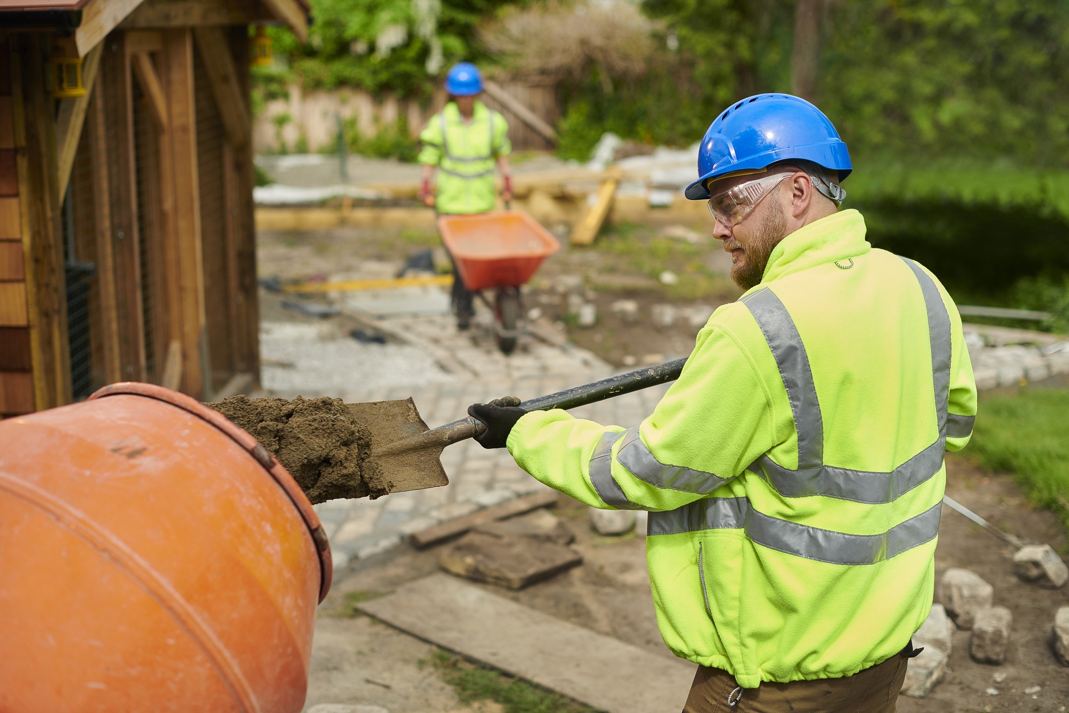 Two construction workers in high-visibility jackets and helmets are engaged in outdoor work, using a mixing machine and wheelbarrow near a building site.