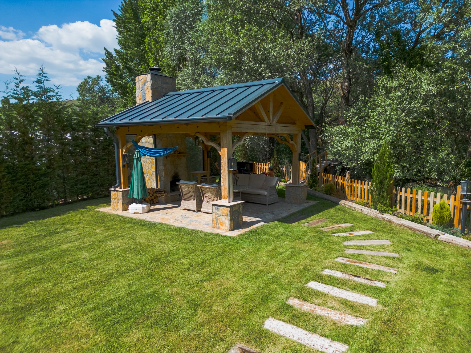 A cozy backyard pavilion with a stone fireplace, surrounded by trees and a fence, features outdoor furniture and a tiled pathway.
