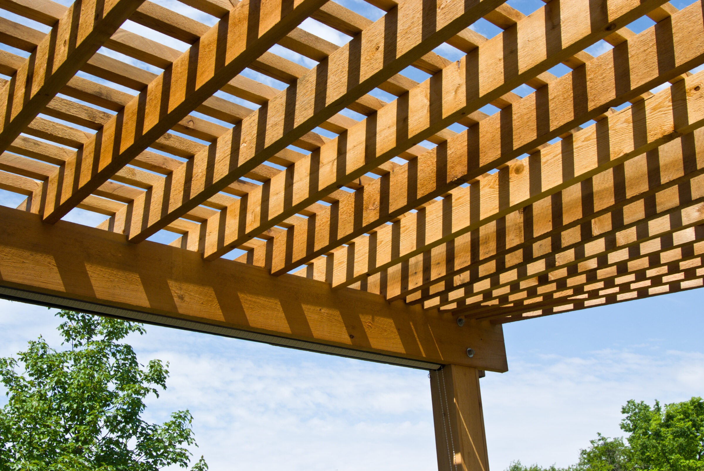 Wooden pergola casting geometric shadows under clear sky, with lush green trees in the background, creating a serene outdoor atmosphere.