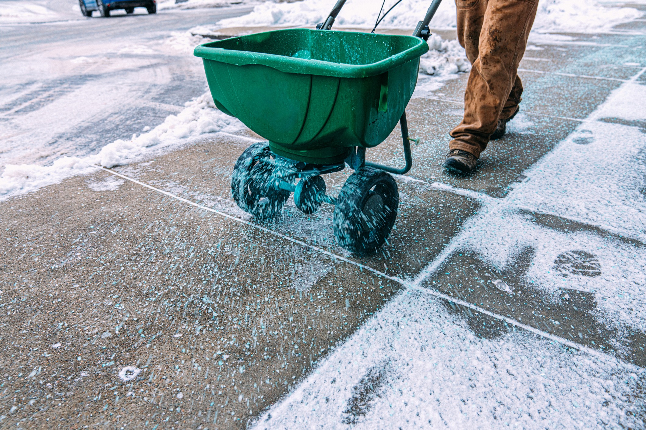 A person spreads de-icing salt on a snowy sidewalk using a green spreader, with snowy ground and parked cars in the background.