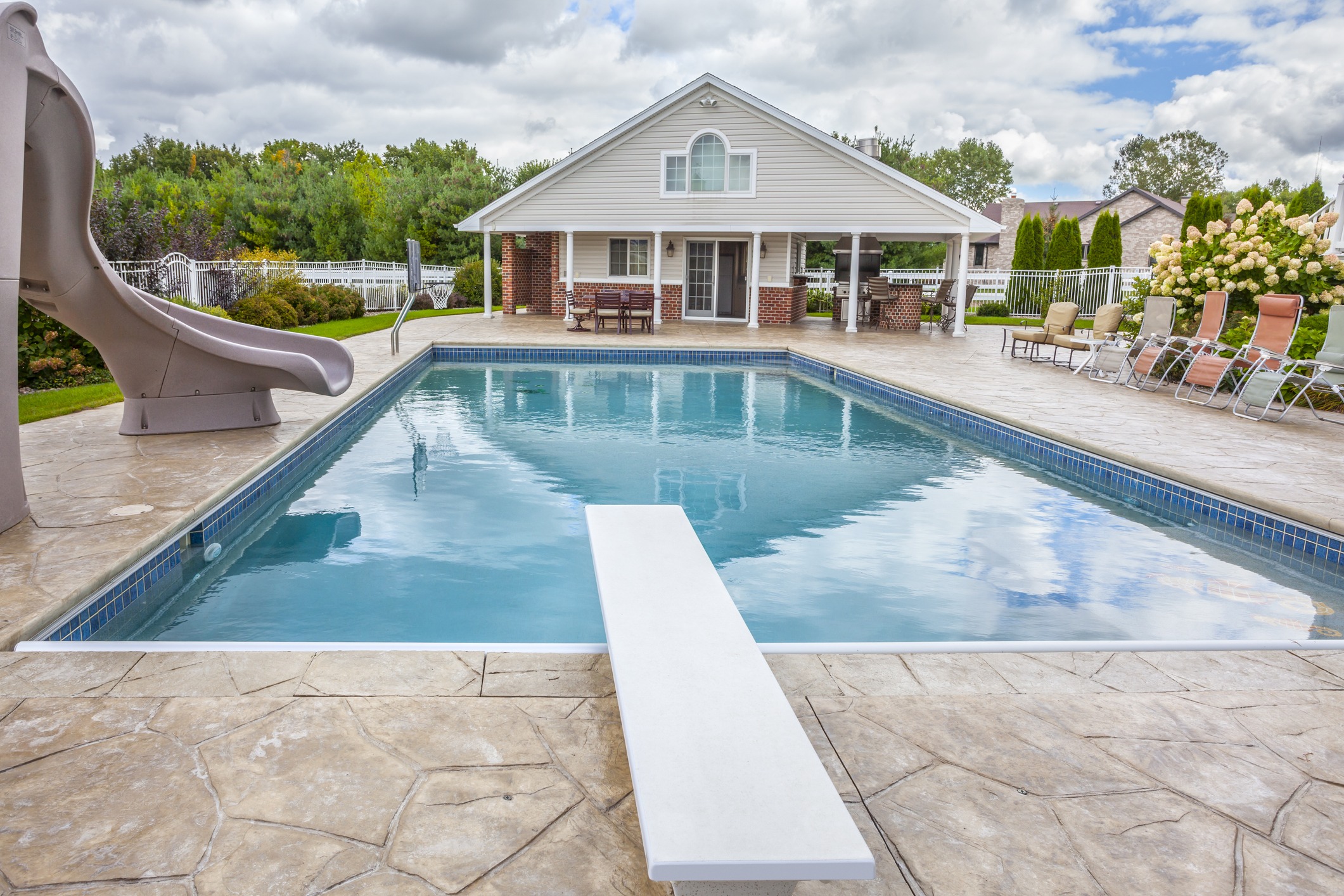 A backyard pool scene with a diving board, slide, patio seating, and a house. Surrounded by trees and partly cloudy sky.