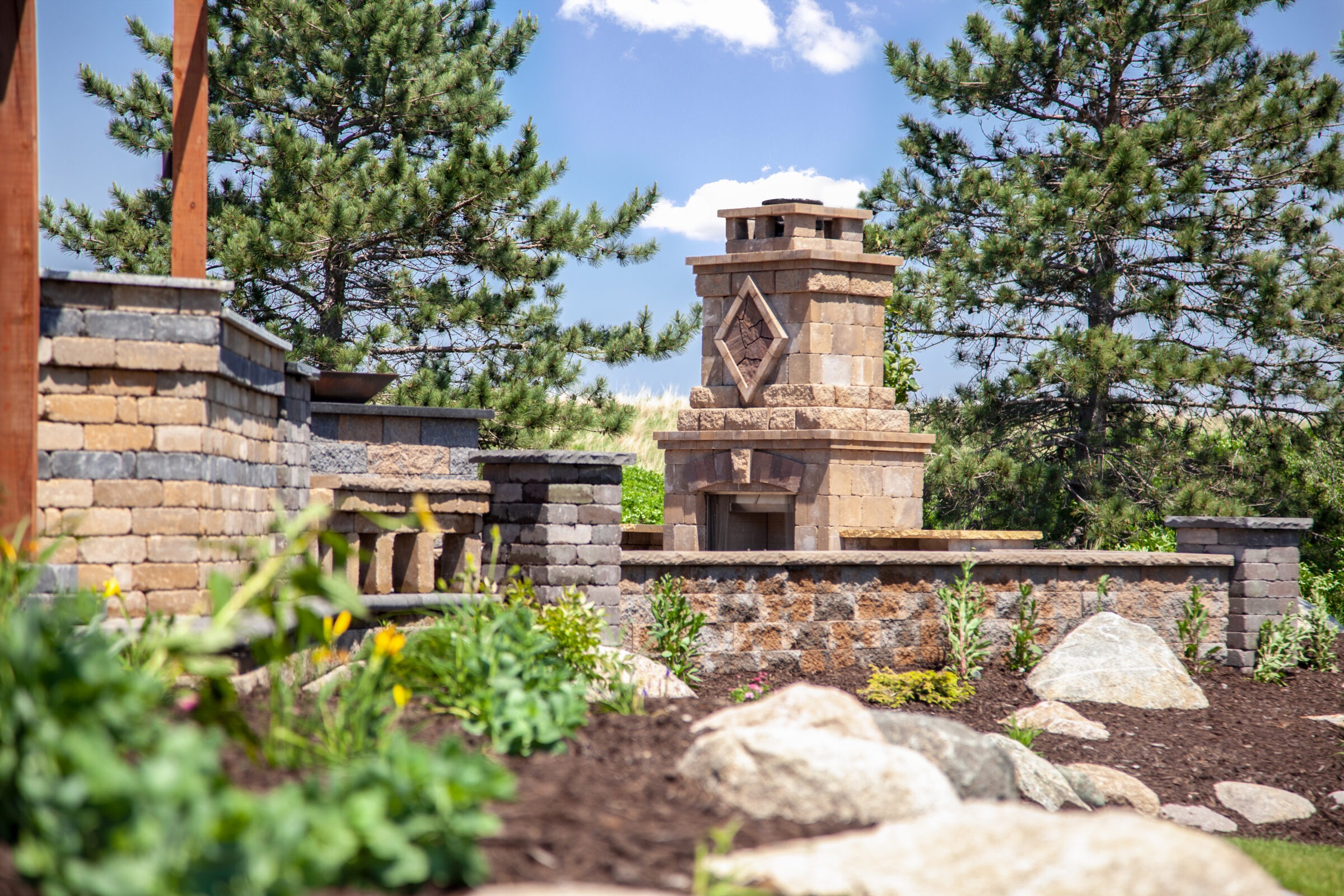 Stone outdoor fireplace surrounded by landscaped garden beds, retaining walls, and pine trees under a blue sky.