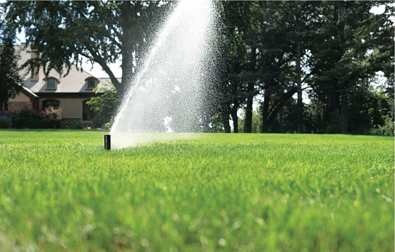 Automatic lawn sprinkler spraying water on a green, well-maintained lawn with a house and trees in the background.