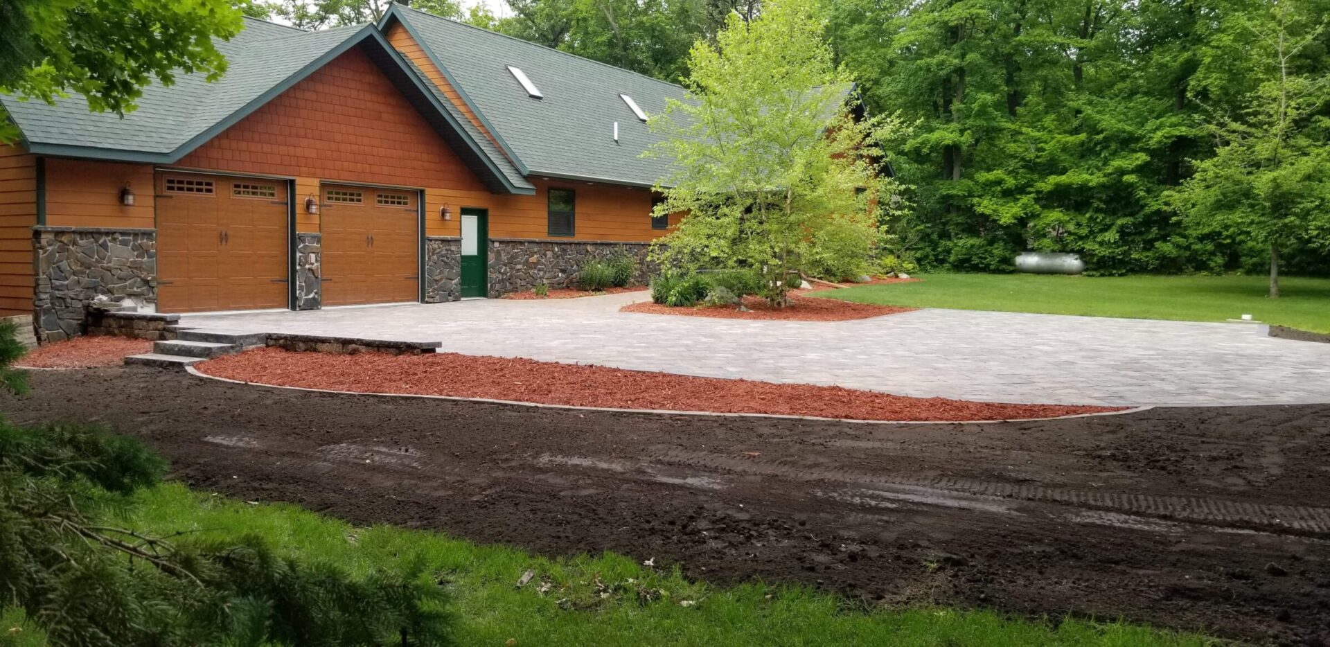 Newly installed paver driveway and mulch landscaping in front of a rustic-style home with stone accents and green roof.